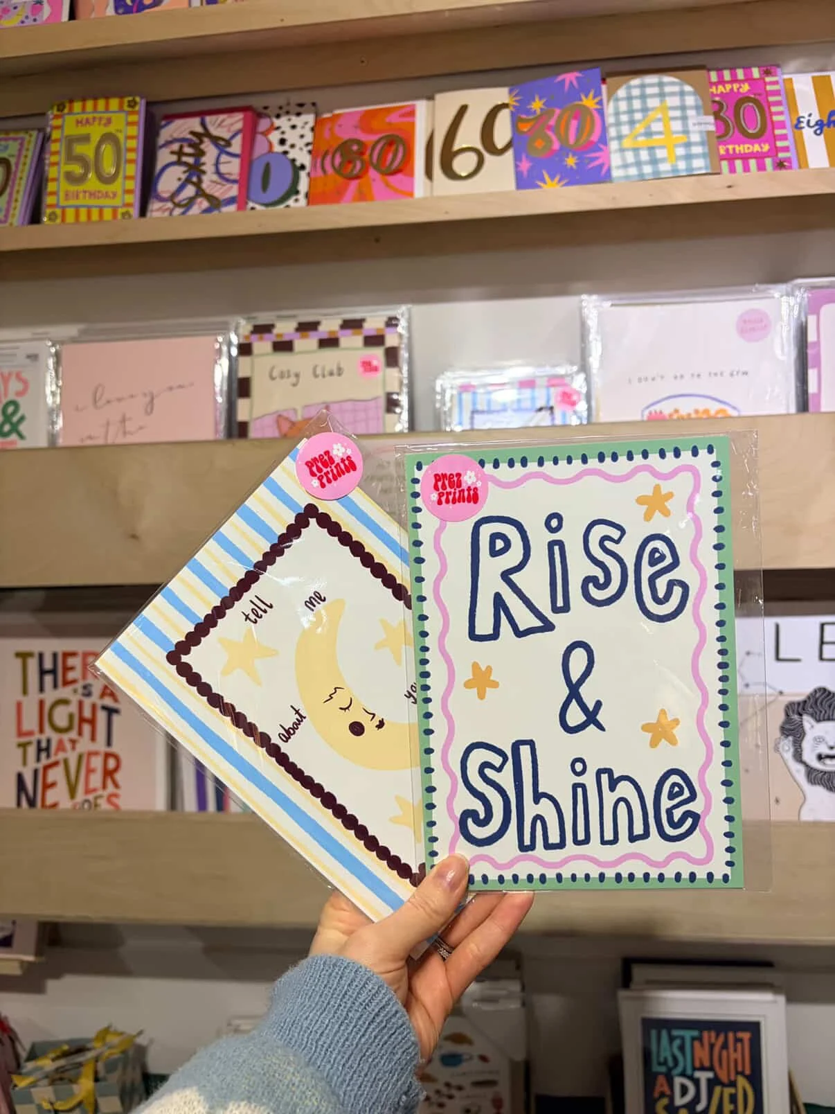 Hand holding two colorful cards, one with the words 'Rise & Shine' surrounded by small stars, and the other with a crescent moon and the phrase 'tell me about you' on a light background with blue and yellow stripes, in a store with other greeting cards on shelves.