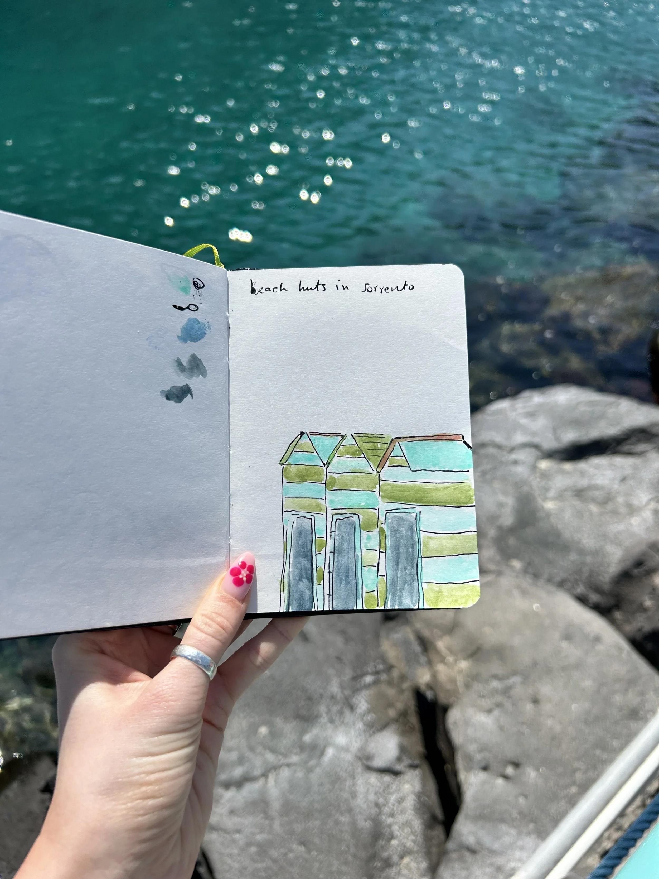 Hand holding a sketchbook with a drawing of colorful beach huts, near rocky shoreline and turquoise water.