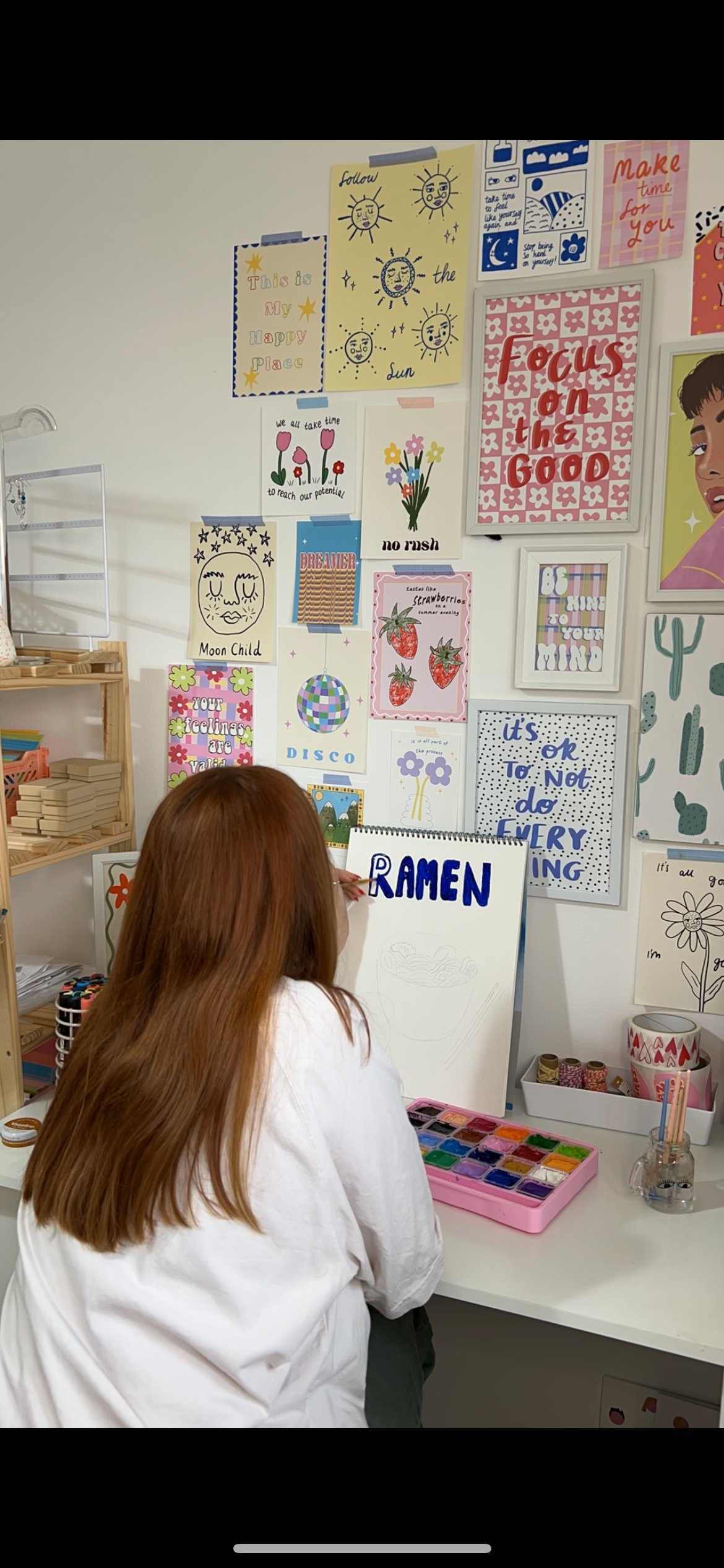 A woman with long reddish hair painting the word 'RAMEN' on a white canvas with blue letters in a colorful art studio decorated with various framed illustrations and motivational quotes.