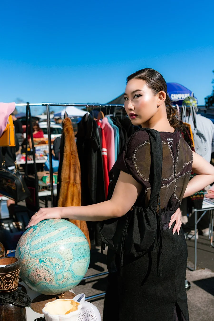 A woman at an outdoor market stands next to a globe, with clothing racks and people shopping behind her on a bright day.
