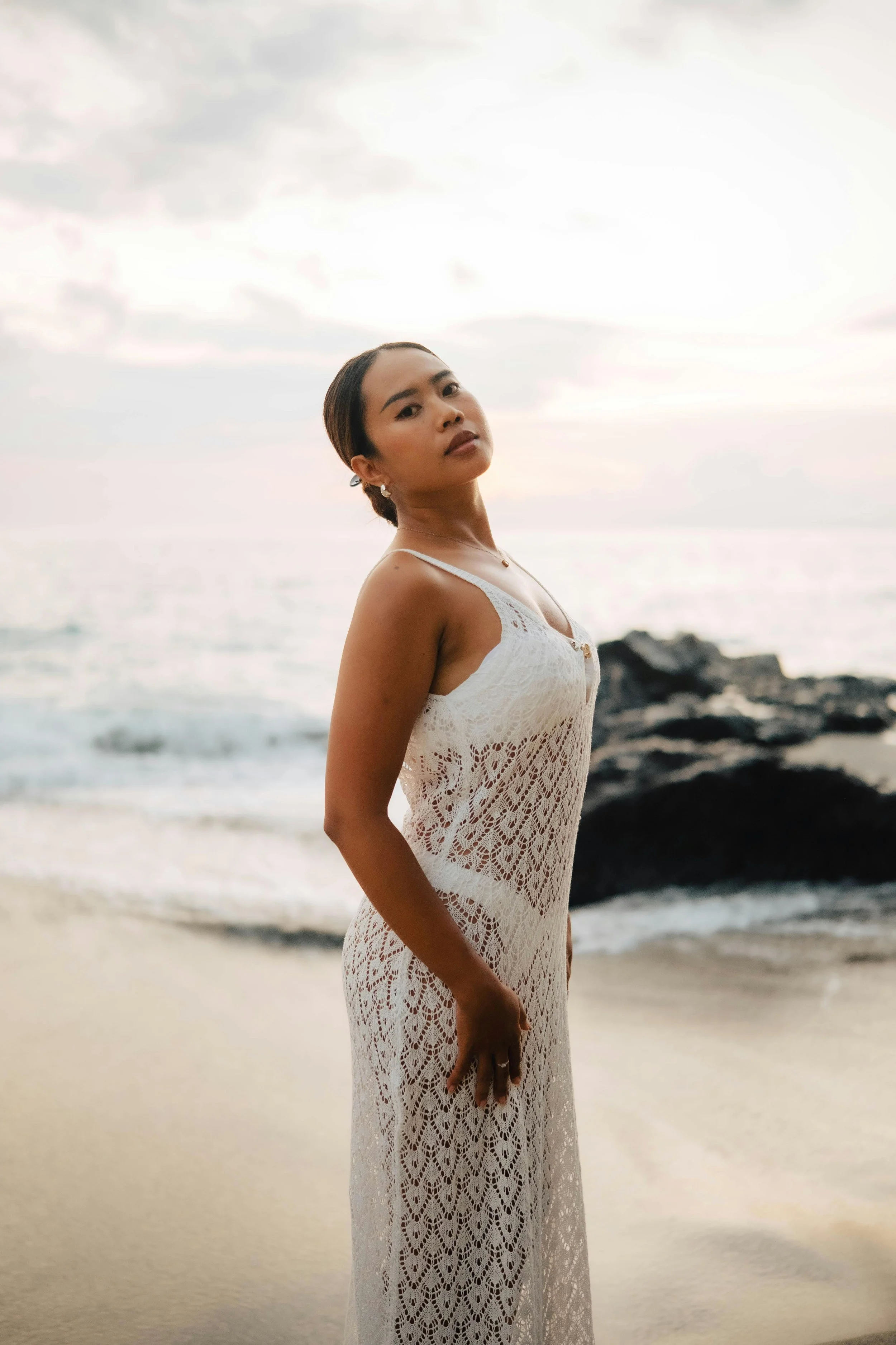 A woman in a white crochet dress standing on a beach with rocks and ocean in the background during sunset.