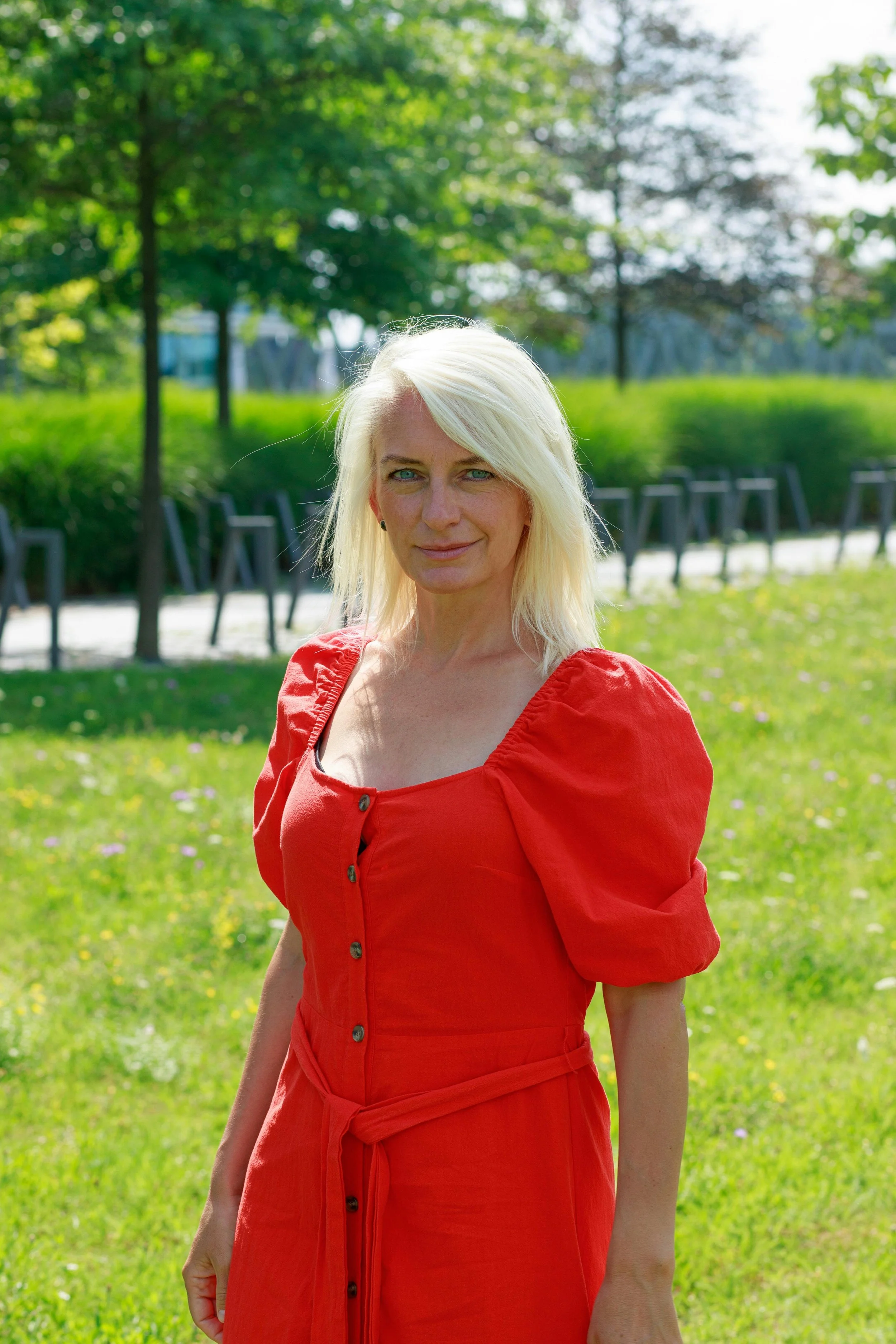 A woman with blonde hair and blue eyes standing outdoors in a park with green trees and grass, wearing a red dress with puffed sleeves.