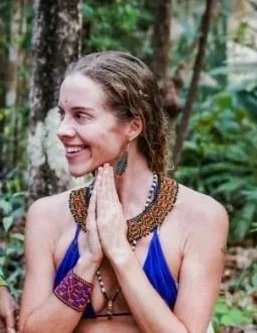 Woman with a joyful smile wearing a blue top, jewelry, and folded hands in a forest setting.