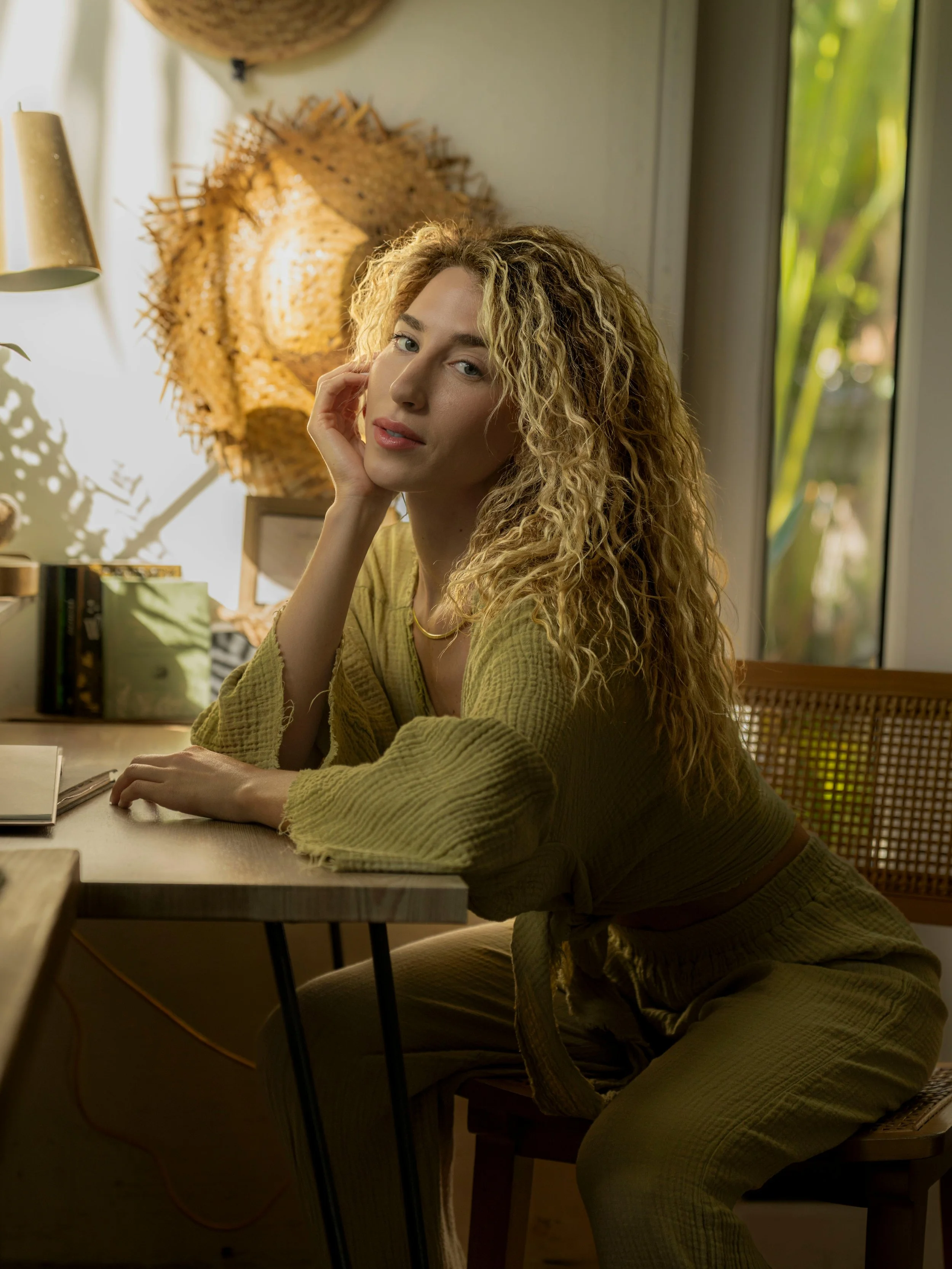 A woman with curly blonde hair, wearing a yellow outfit, sitting at a wooden desk in a well-lit room with large windows.