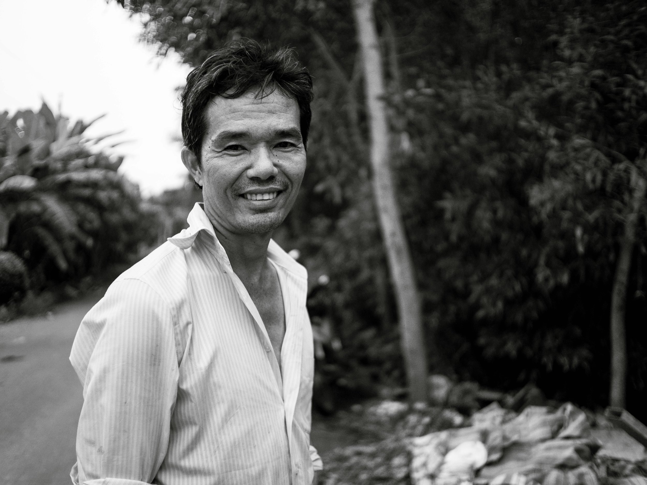 A smiling man standing outdoors, wearing a light-colored, collared, striped shirt, with trees and foliage in the background.