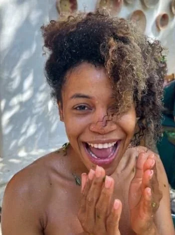 A woman with curly hair smiling and clapping in front of a backdrop with circular patterns.
