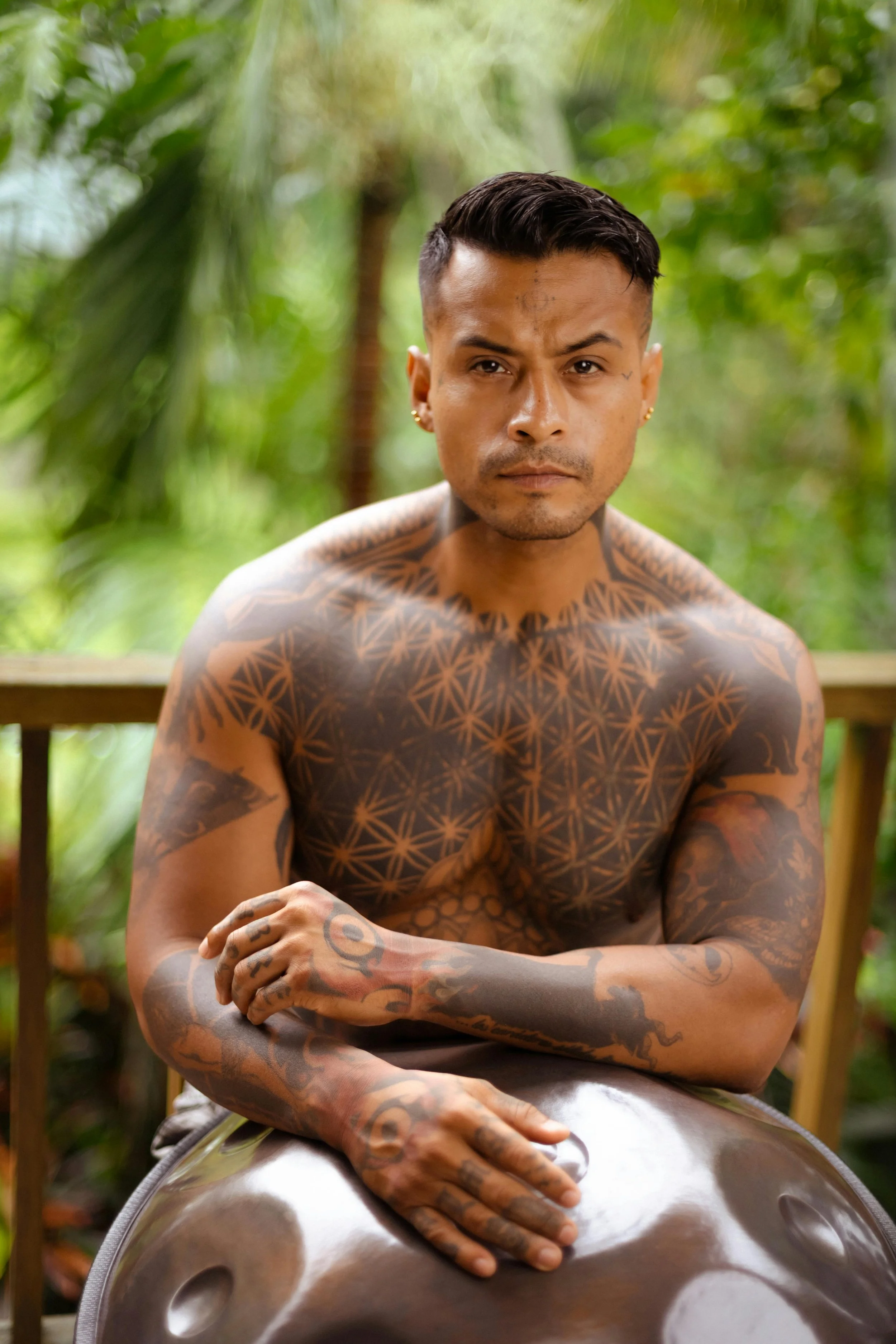 A shirtless man with numerous tattoos sitting outdoors in a lush, green environment, playing a steel tongue drum.