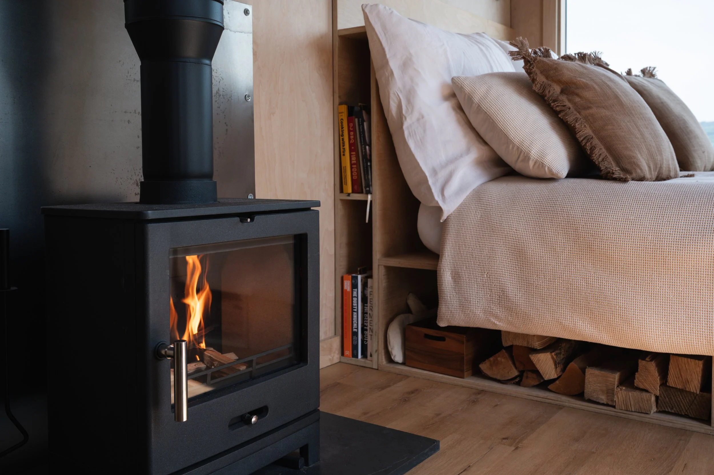 Cozy tiny house interior featuring a modern wood-burning stove next to a comfortable bed with neutral linens and rustic wood storage.