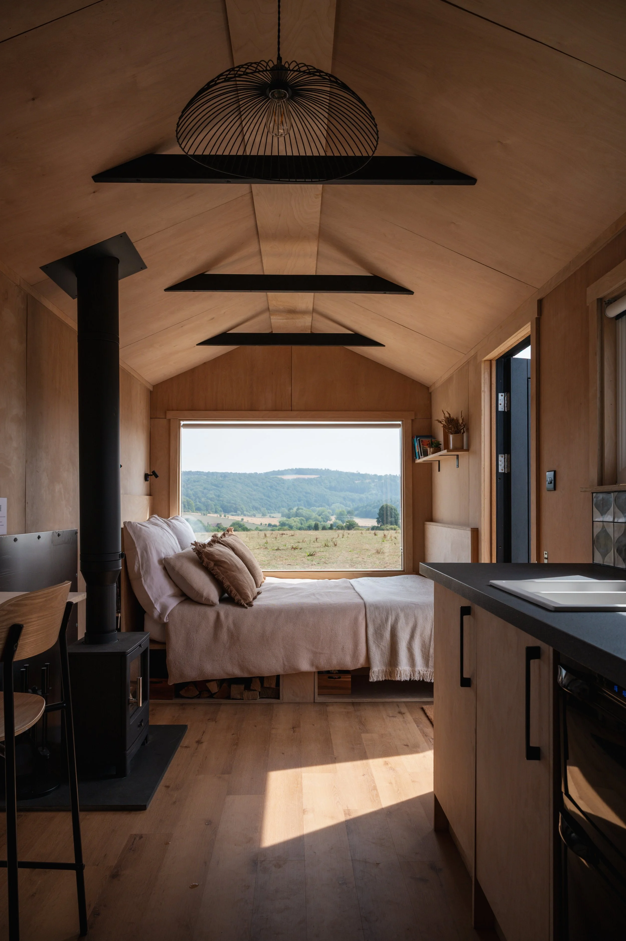 Tiny house interior with wood-burning stove, kitchen, and bed facing panoramic countryside views through a large window.