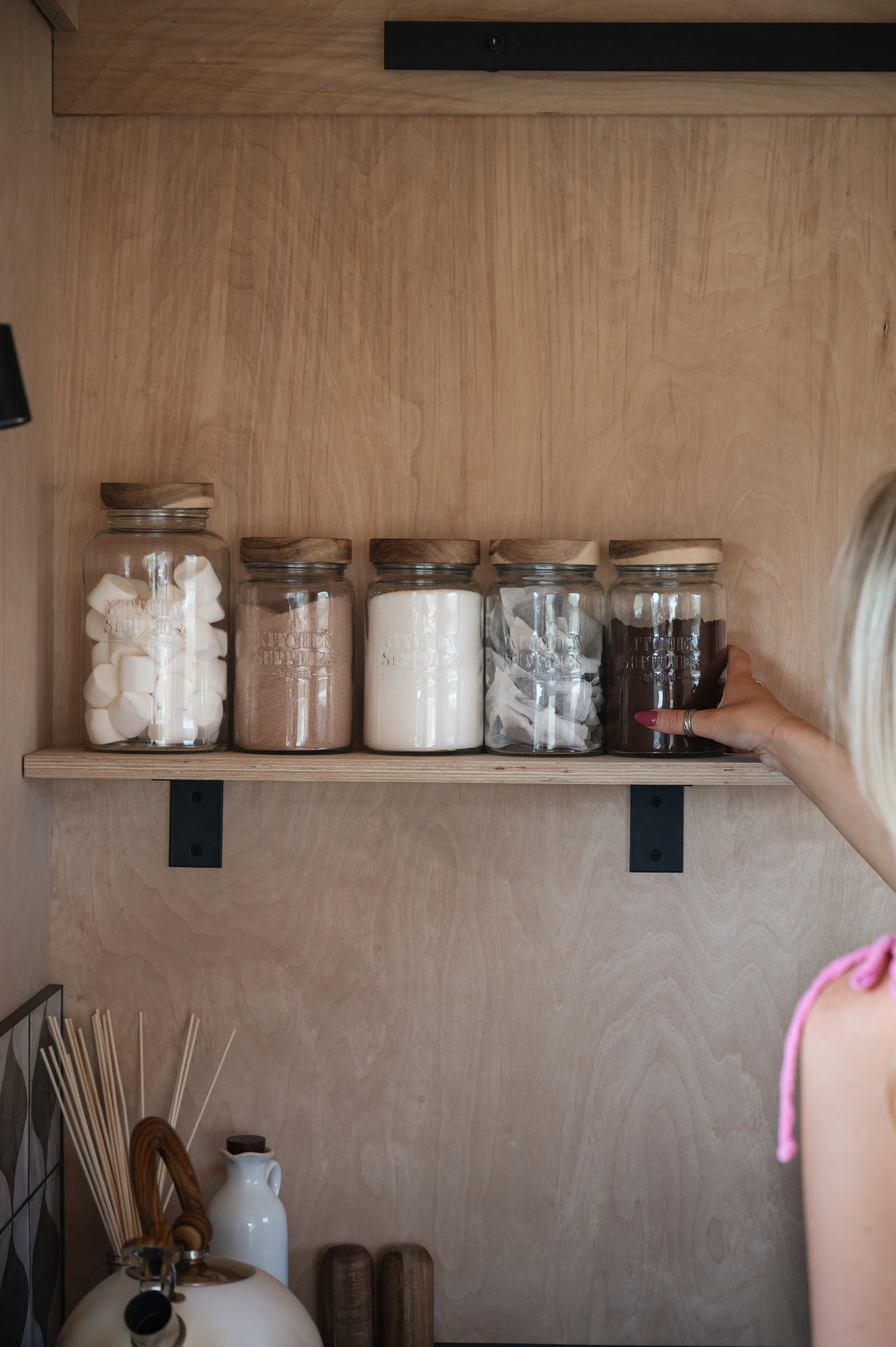 Tiny house kitchen shelf with glass jars of hot drinks and treats — marshmallows, cocoa, sugar, tea, and coffee storage.