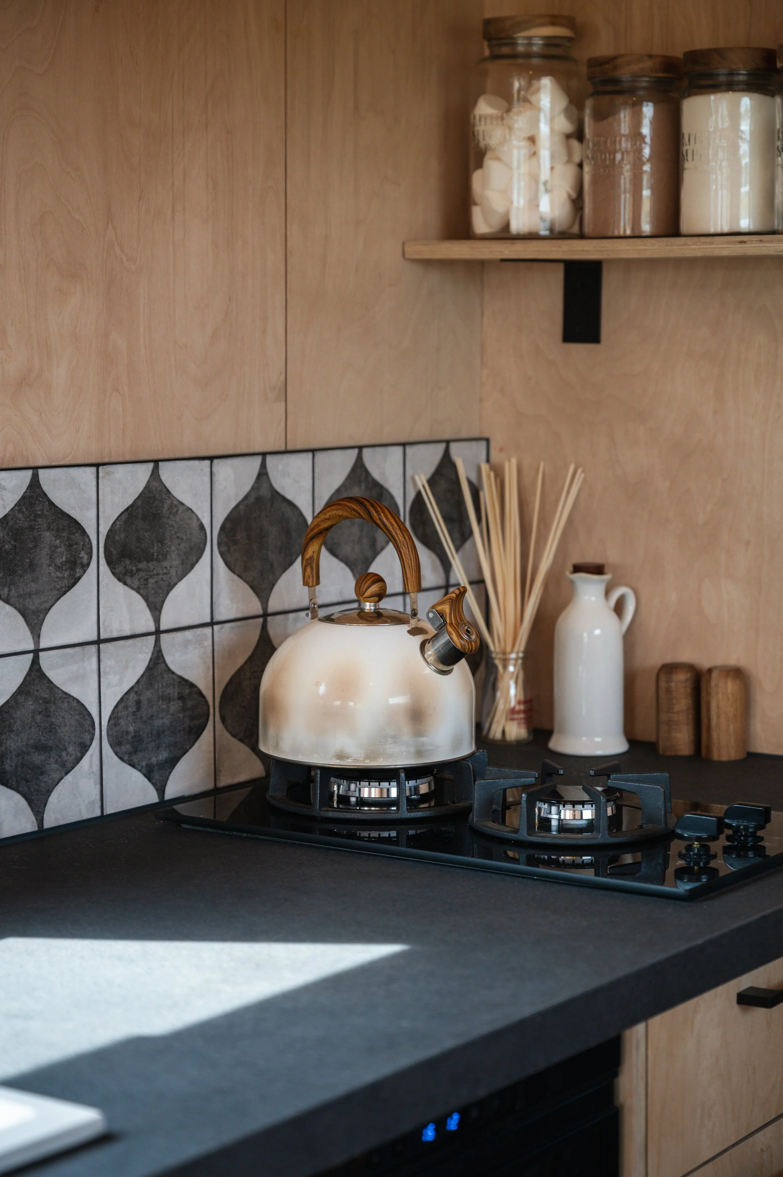 Charming tiny house kitchen design featuring a gas hob with rustic kettle, modern patterned tiles, and neatly styled shelves with glass jars for a cosy, functional space.