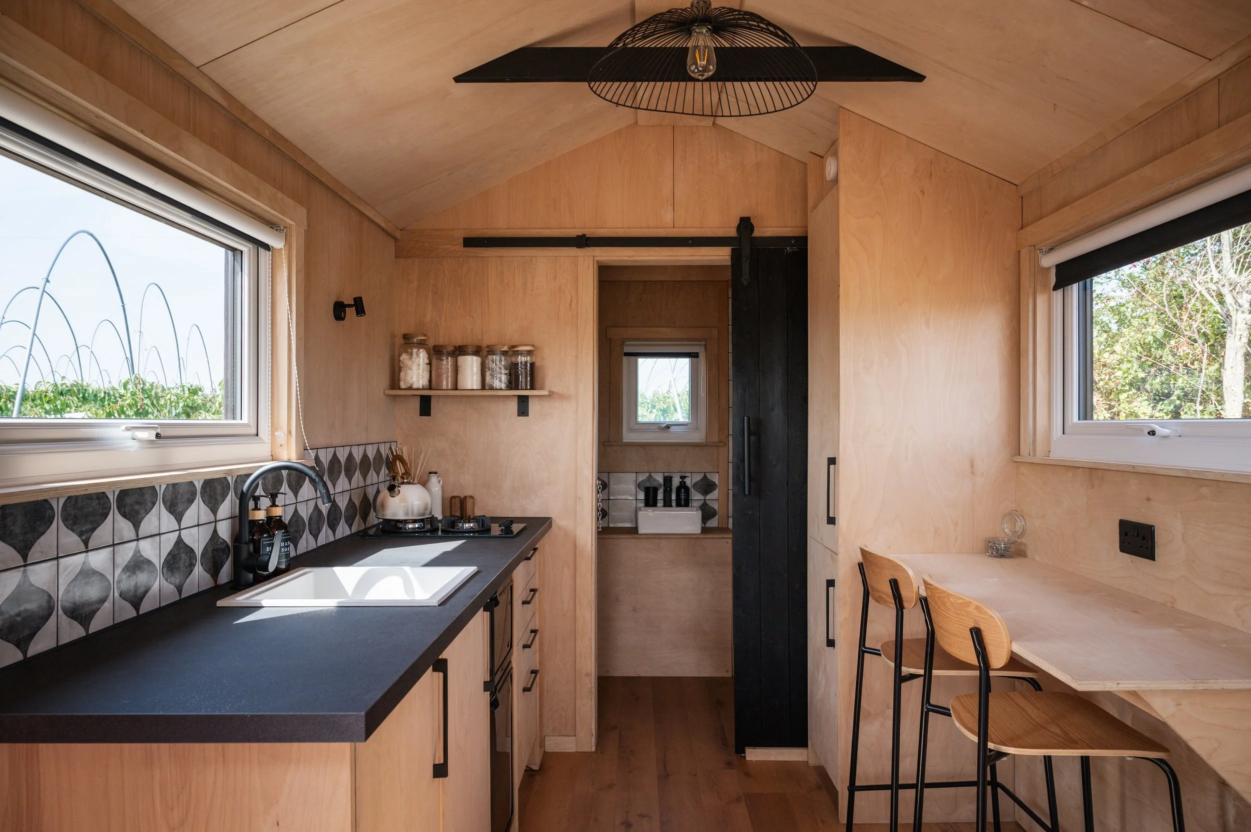 Tiny house kitchen and dining area with black countertop, gas hob, compact breakfast bar, and Beech plywood interior — off-grid cabin design.
