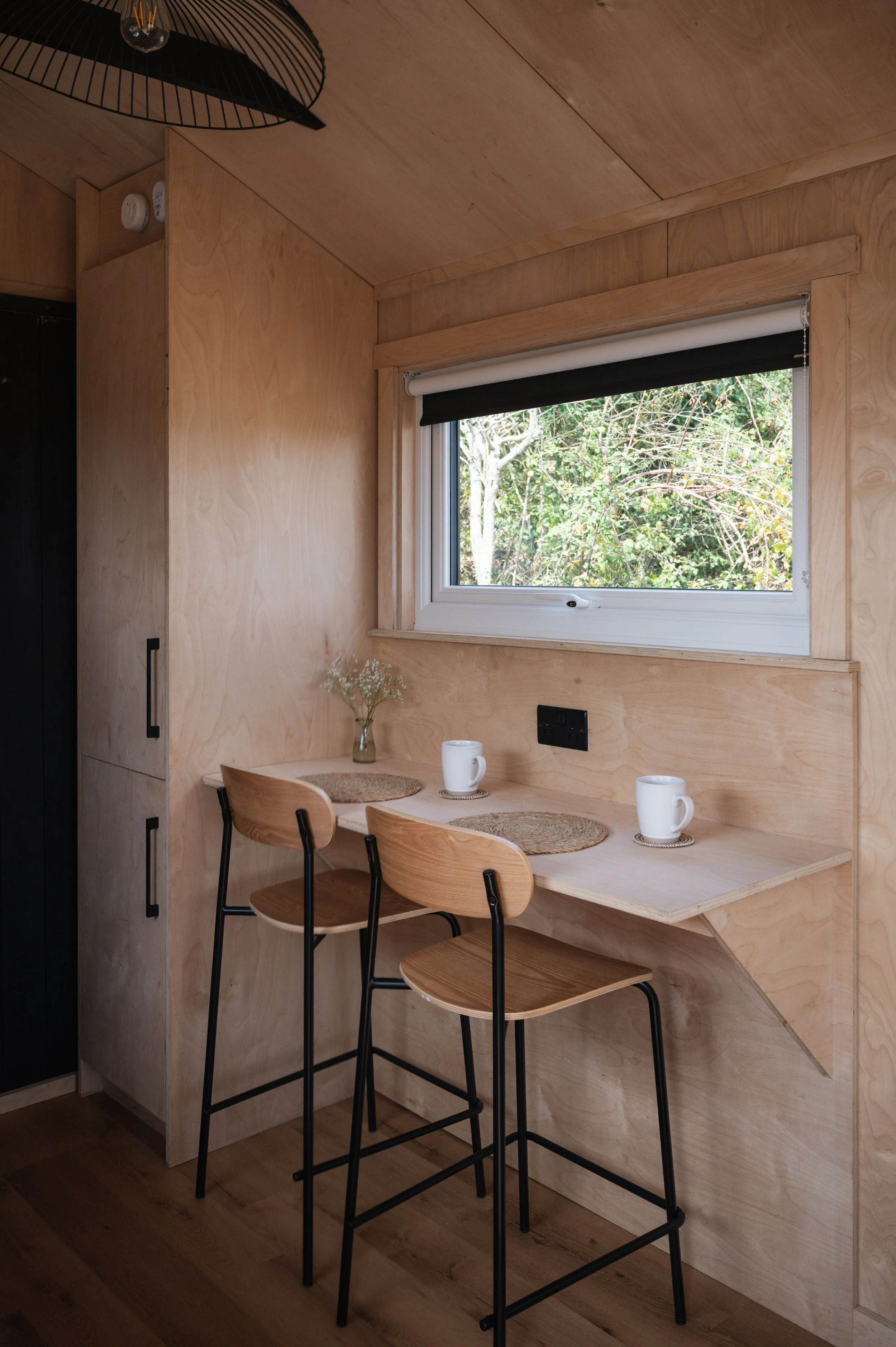 Compact dining area inside a tiny home with two wooden stools at a built-in table beneath a window, finished with light beech plywood walls and natural décor.