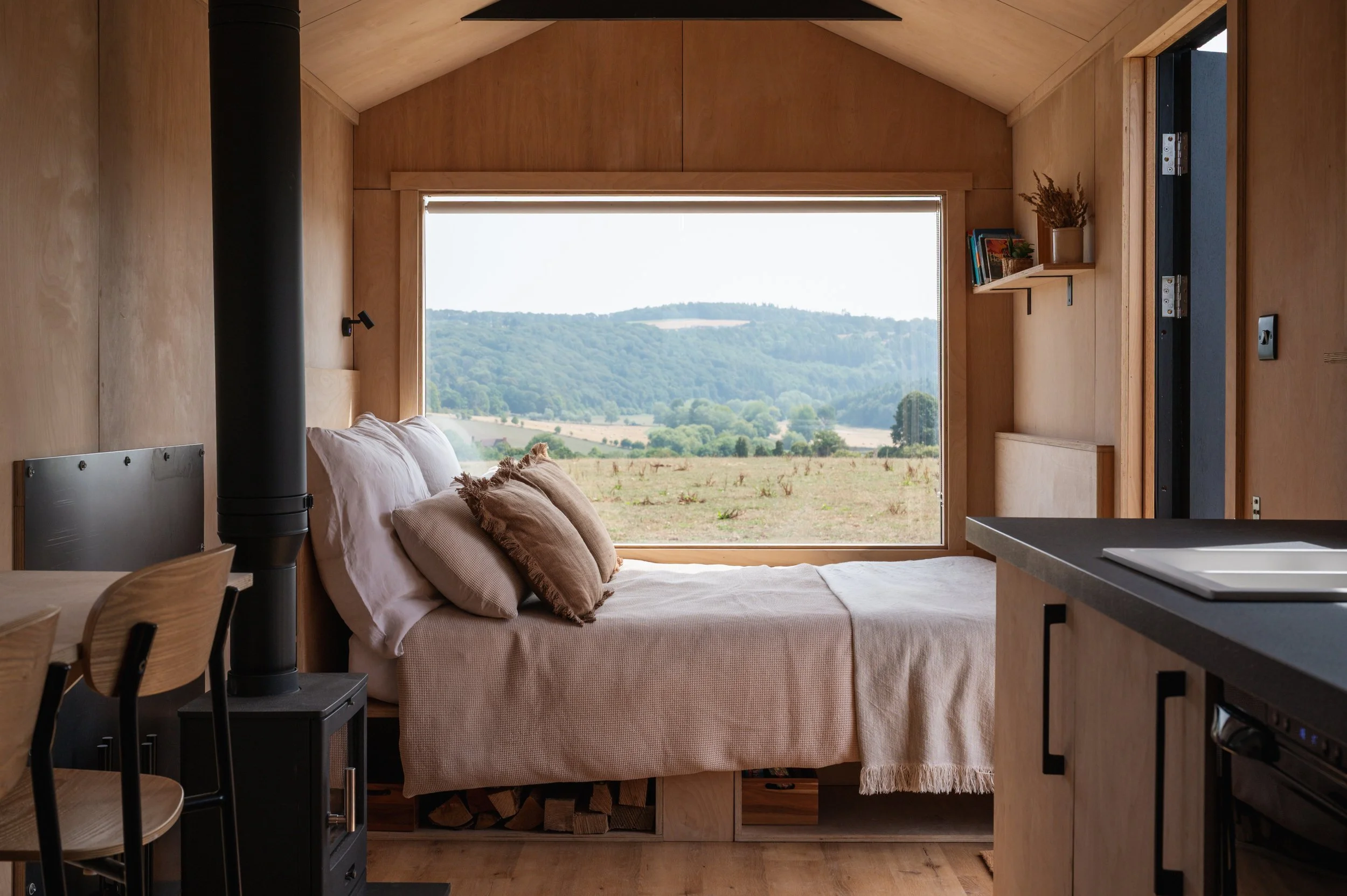 Relax in a modern tiny house with panoramic countryside views from the bed. This eco-friendly cabin features natural wood interiors, a log burner, and a large picture window — the perfect off-grid retreat.
