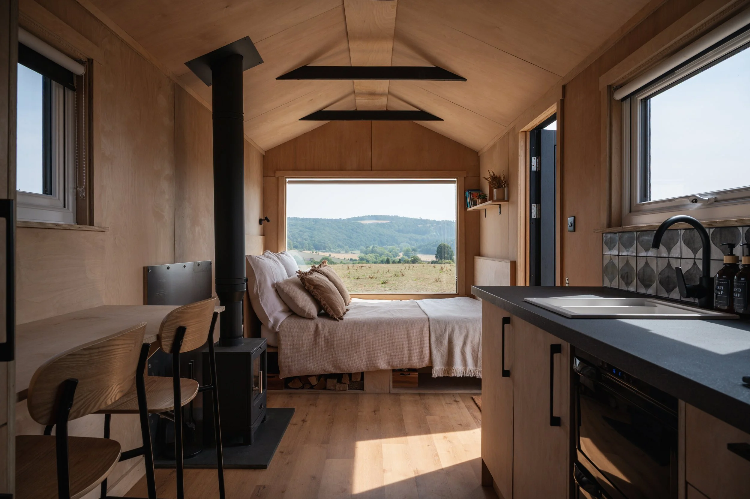 Tiny house interior with wood-burning stove, kitchen, and bed facing panoramic countryside views through a large window.