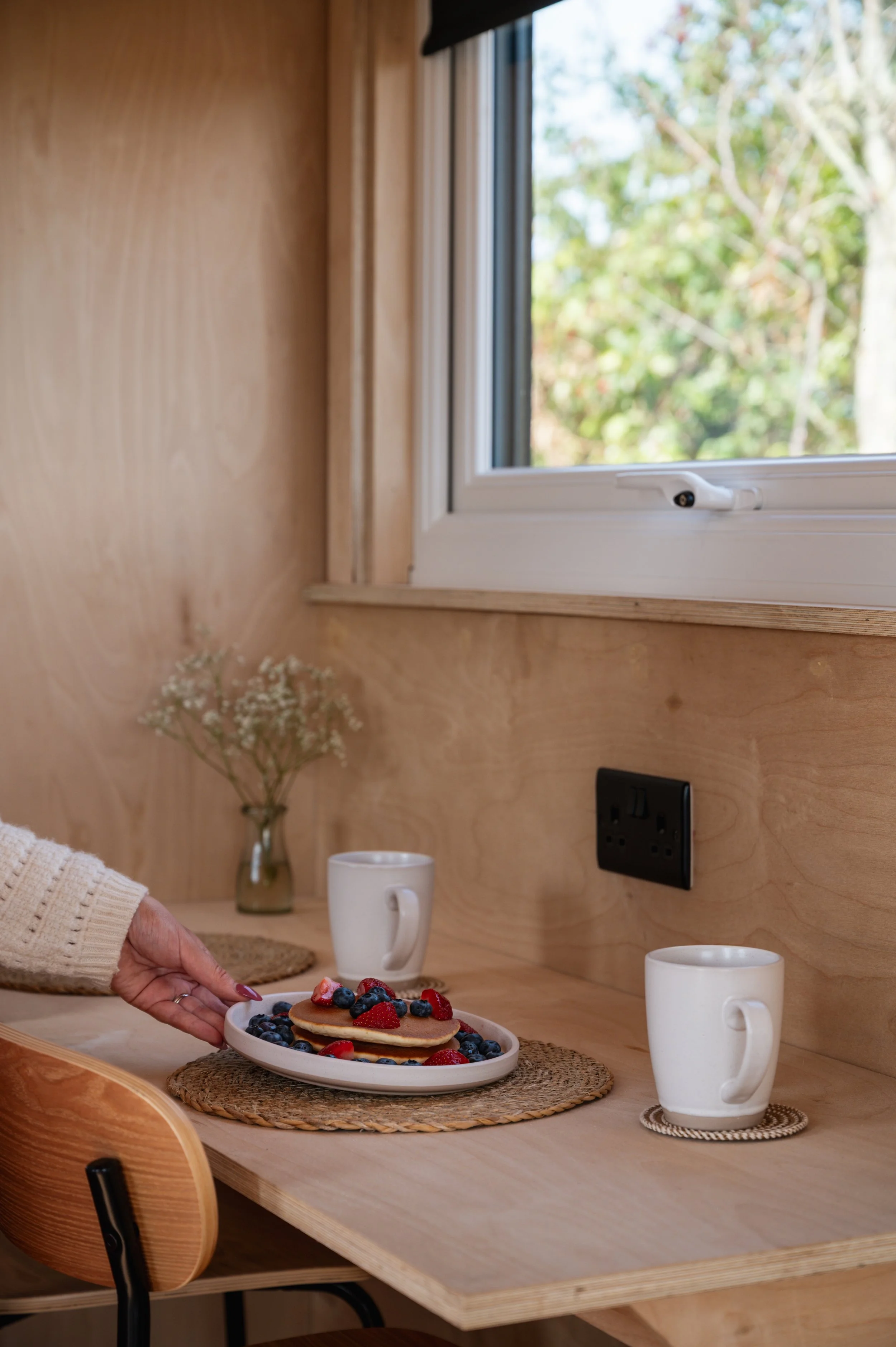 Cozy dining space inside a tiny house, featuring a wooden breakfast bar with coffee mugs and fresh berry pancakes, set beside a window with views of the outdoors.