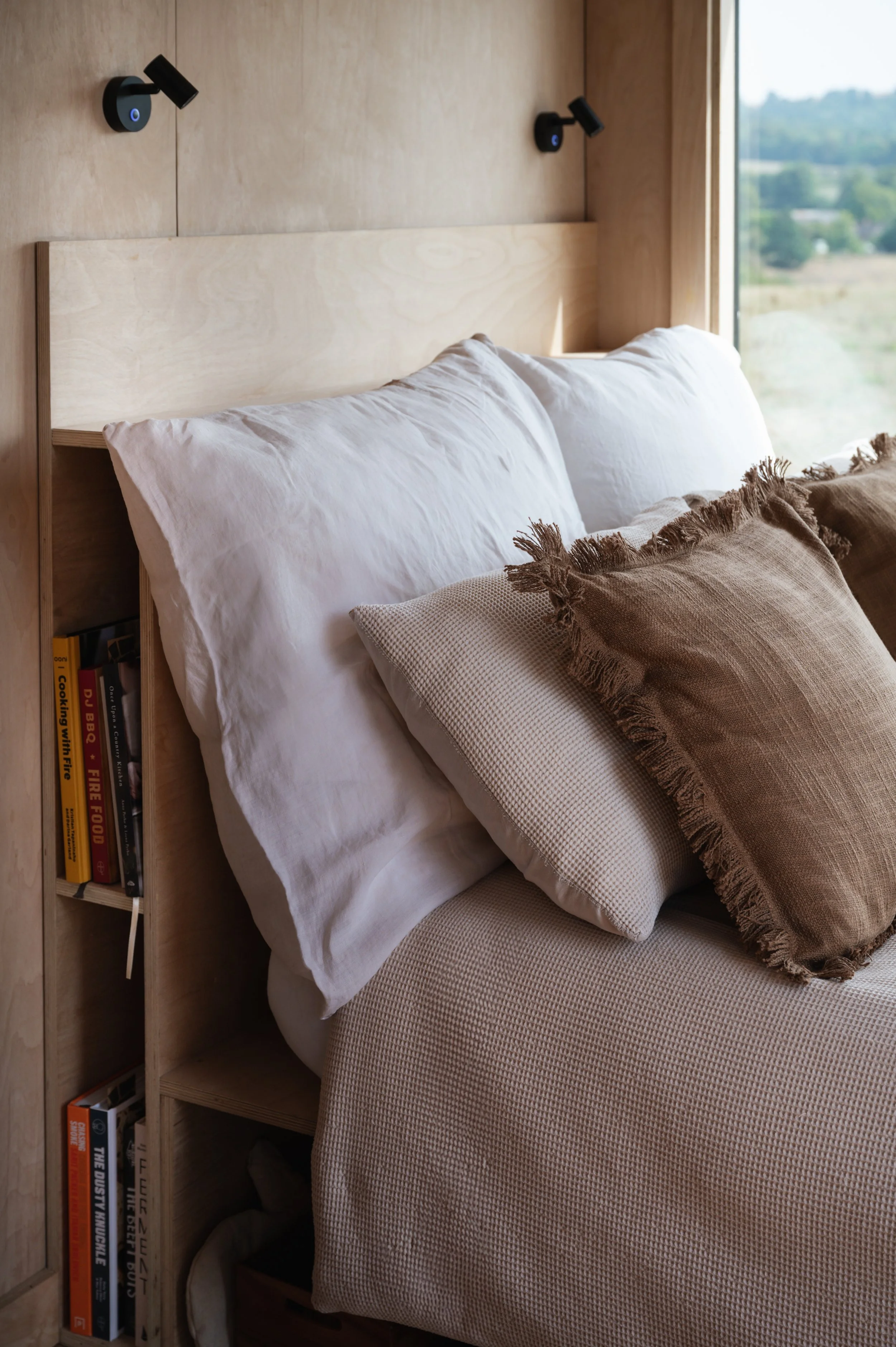Unwind in a tiny house retreat featuring a snug bed with neutral linens, rustic pillows, and a clever built-in bookshelf. Ideal for stays with a touch of homely charm.