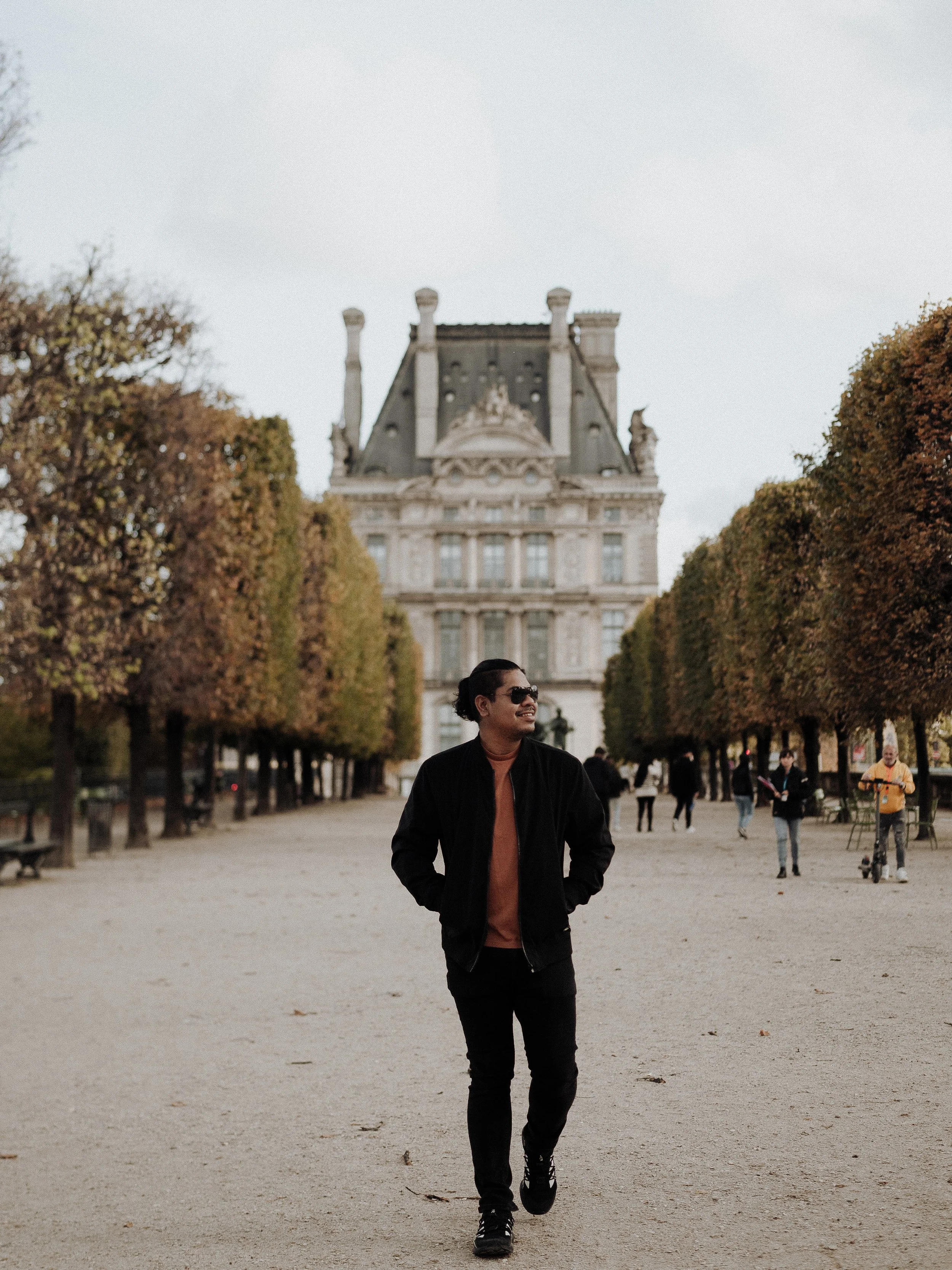 A man wearing sunglasses and a black jacket walking in front of a historic building with a grey slate roof, surrounded by trees with autumn foliage, in a spacious park or courtyard.