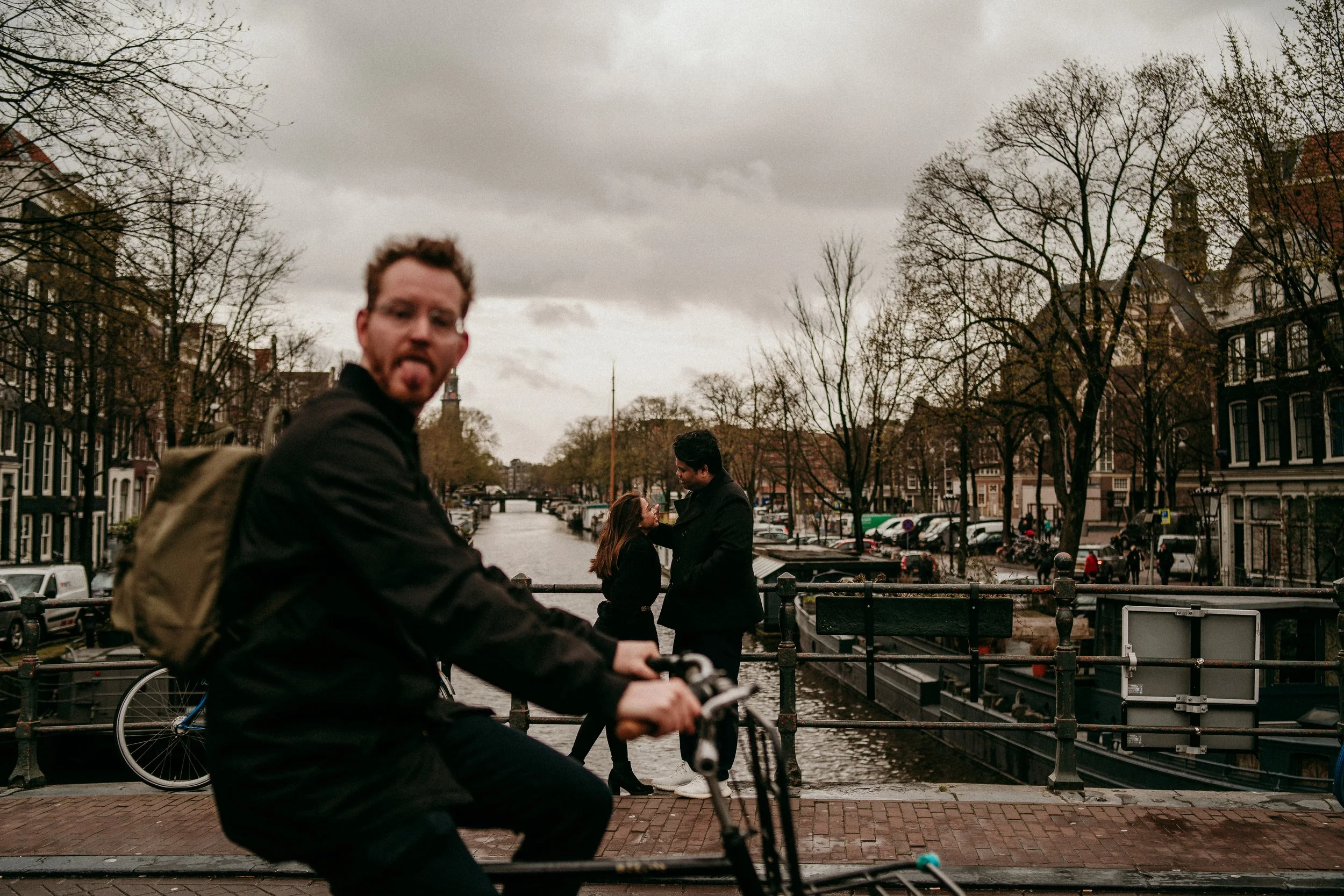 A man riding a bicycle in the foreground with a man and woman in the background standing on a bridge over a canal in a city, with leafless trees, parked cars, and historic buildings visible.