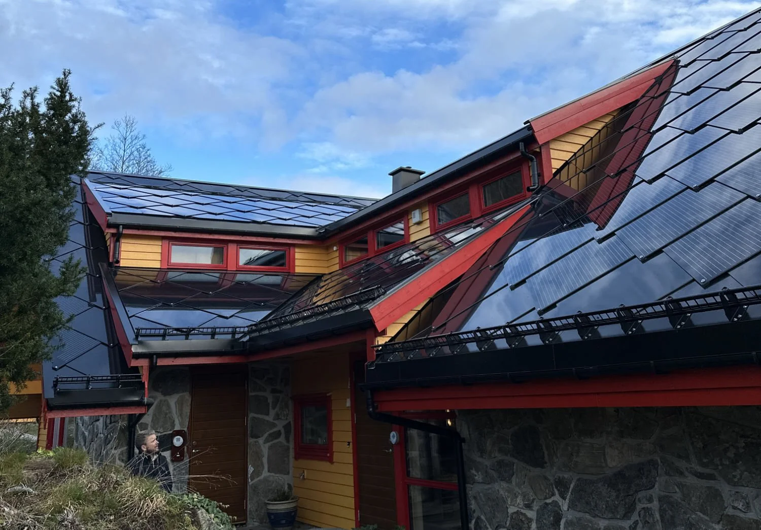 Et hus med moderne design, takdekkende med solcellepaneler, belagt med svart metalltak, vegger av tre og stein, og et tre til venstre i forgrunnen.