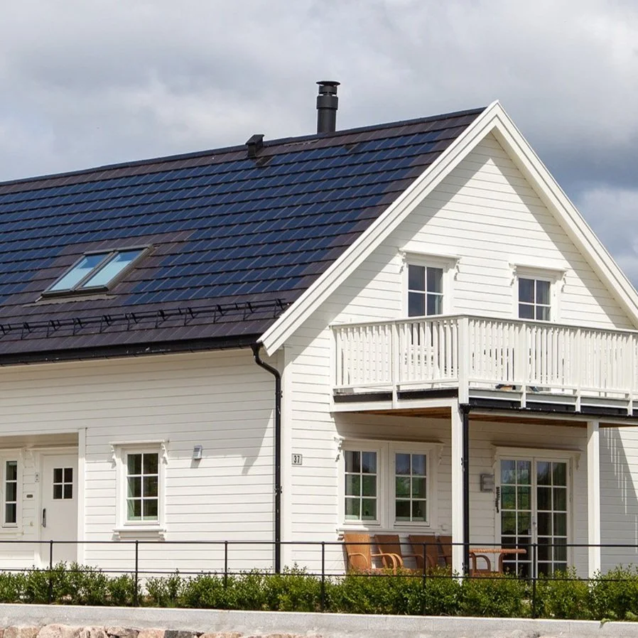 Hus med hvit utvendig panel, svart tak, frontveranda med stoler, og balkong på andre etasje.