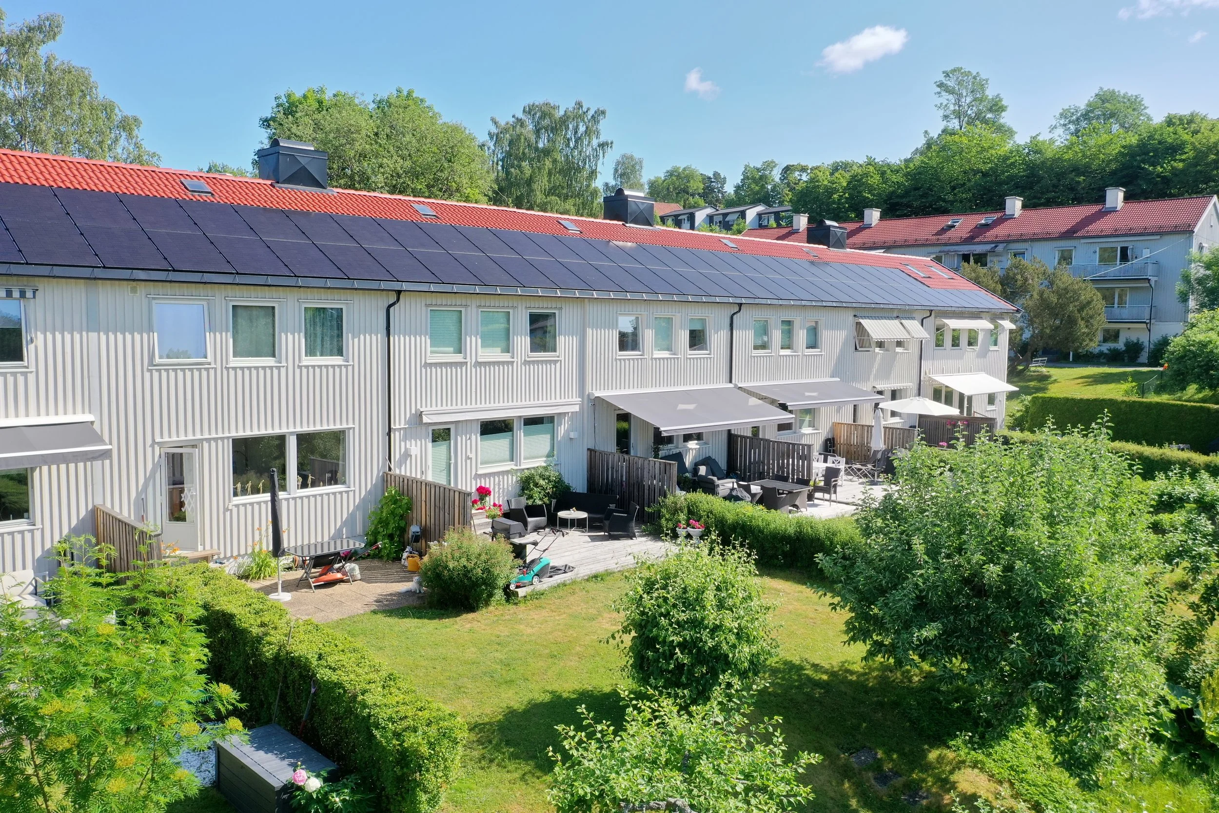 Utsikt over rekkehus med grønne hager, terrasser og solcellepanel på taket, omgitt av trær.