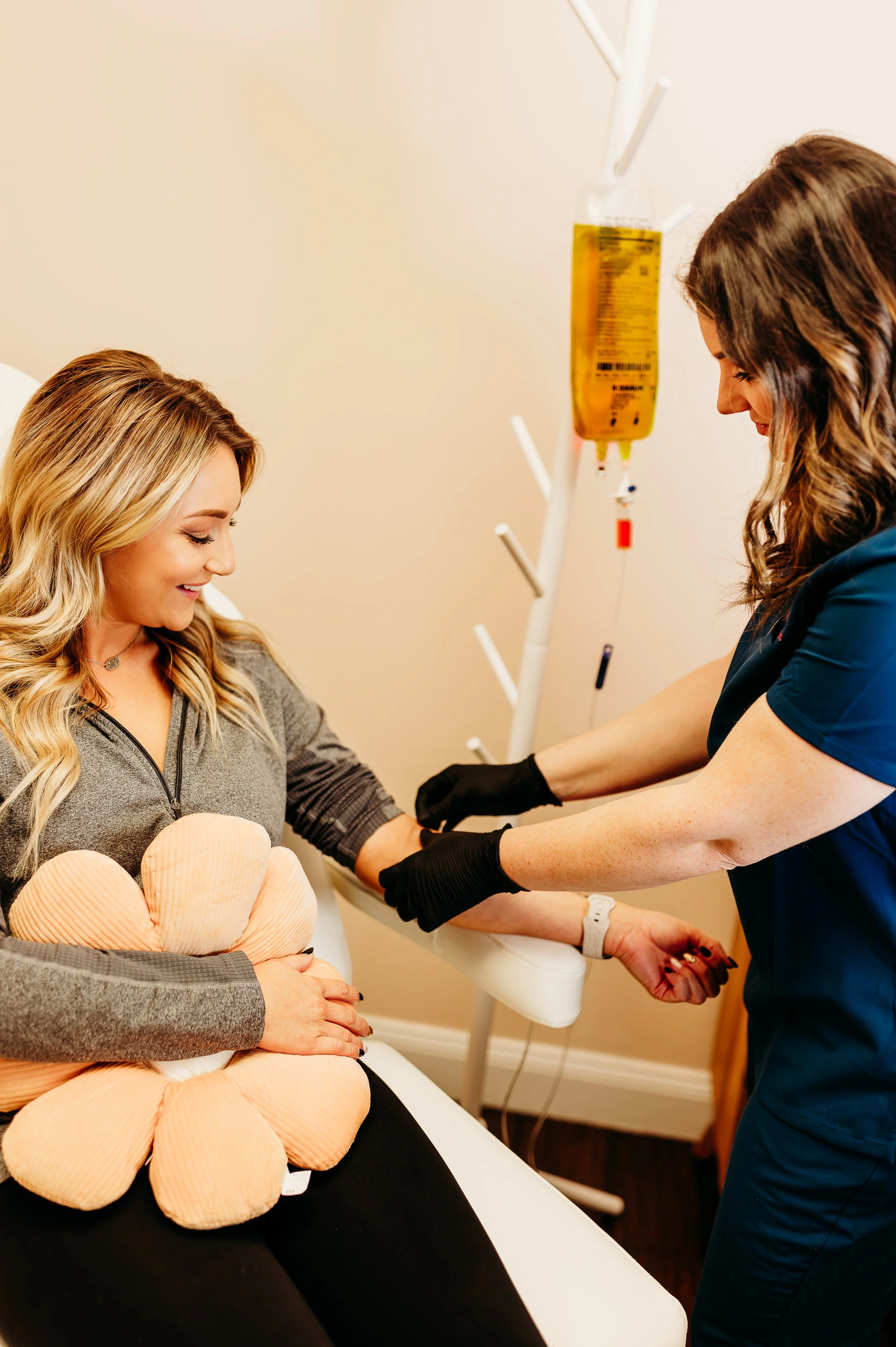 A woman with a teddy bear-shaped pillow on her lap receives a medical injection from a nurse in a clinical setting, with IV fluids hanging on a stand nearby.