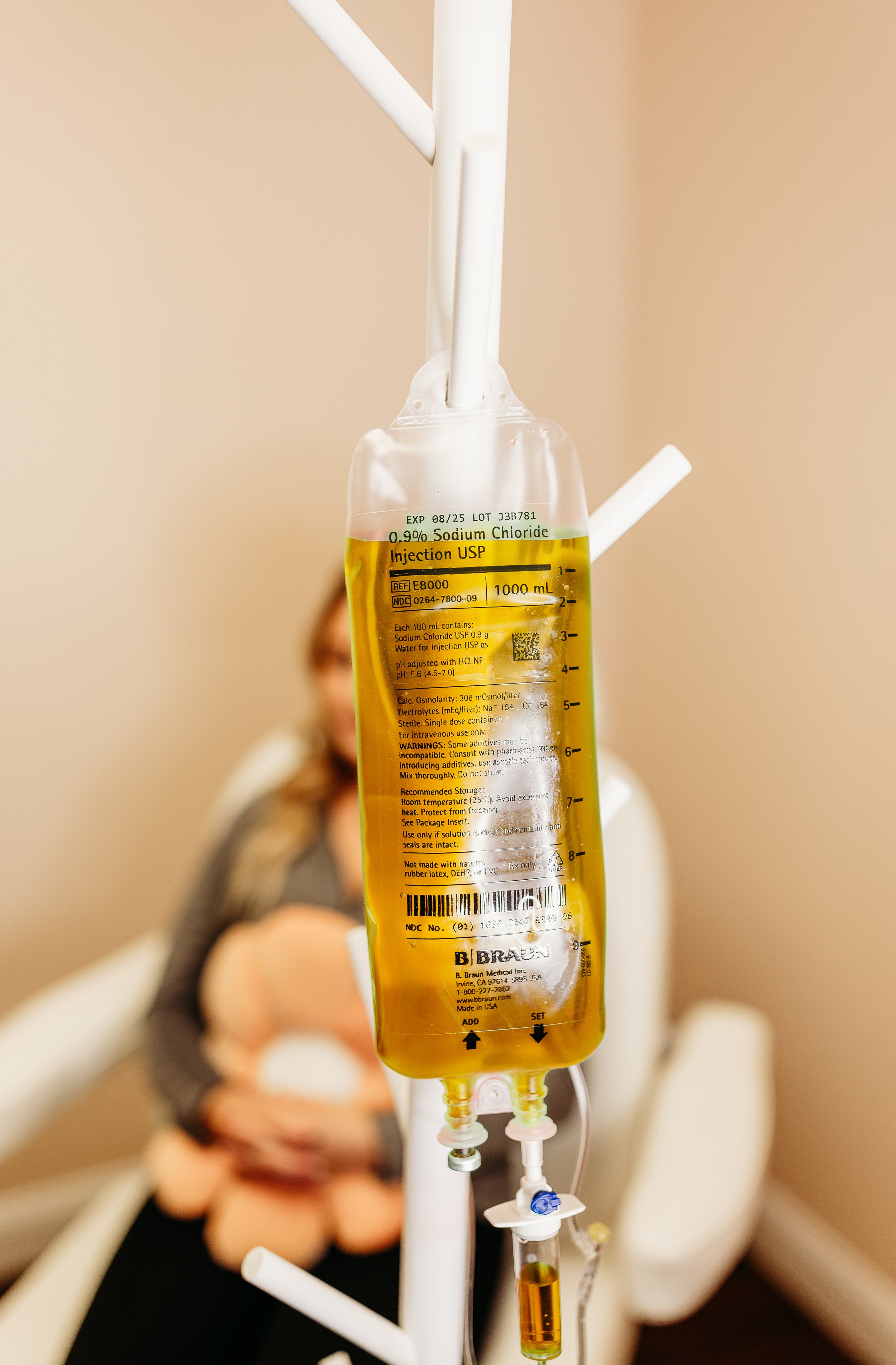 Medical IV drip containing yellowish liquid, hanging in a hospital or clinic room with a blurred patient in the background.