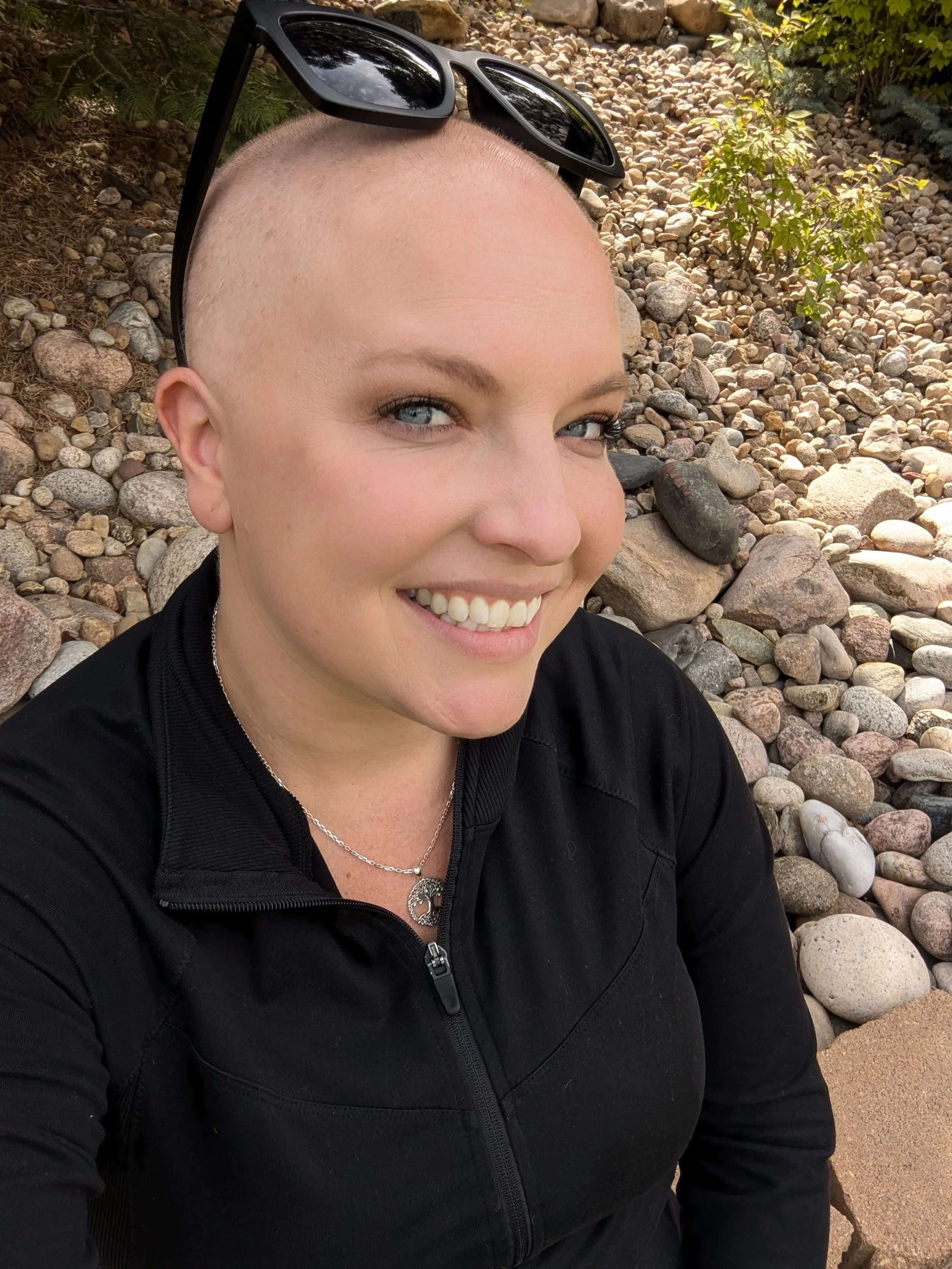 Person with shaved head and sunglasses smiling outdoors in a rocky garden setting.