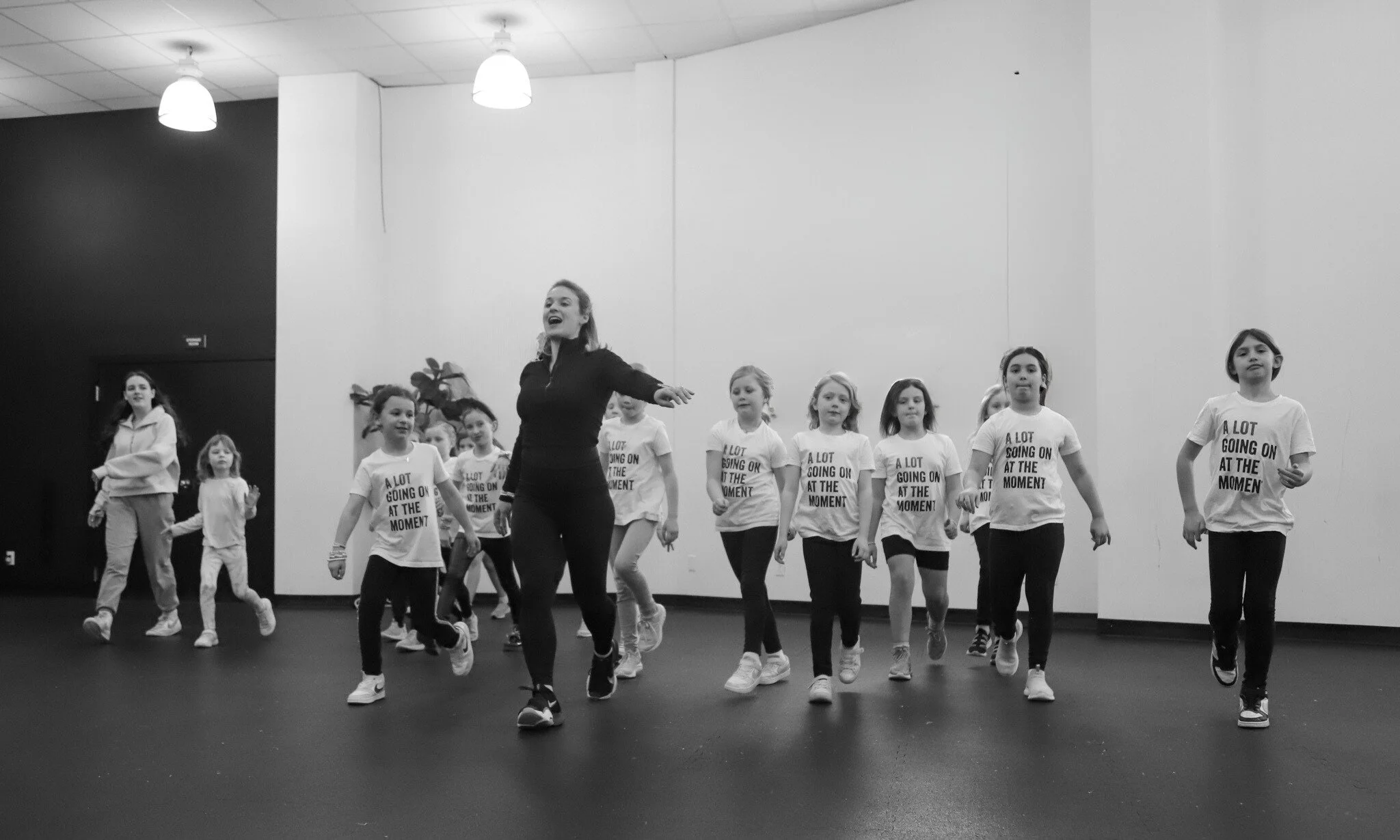 A group of children and an instructor in a dance class. The children wear T-shirts with the phrase "A LOT GOING ON AT THE MOMENT," and the instructor is leading them in a dance routine in a studio with dark flooring and white walls, illuminated by ceiling lights.