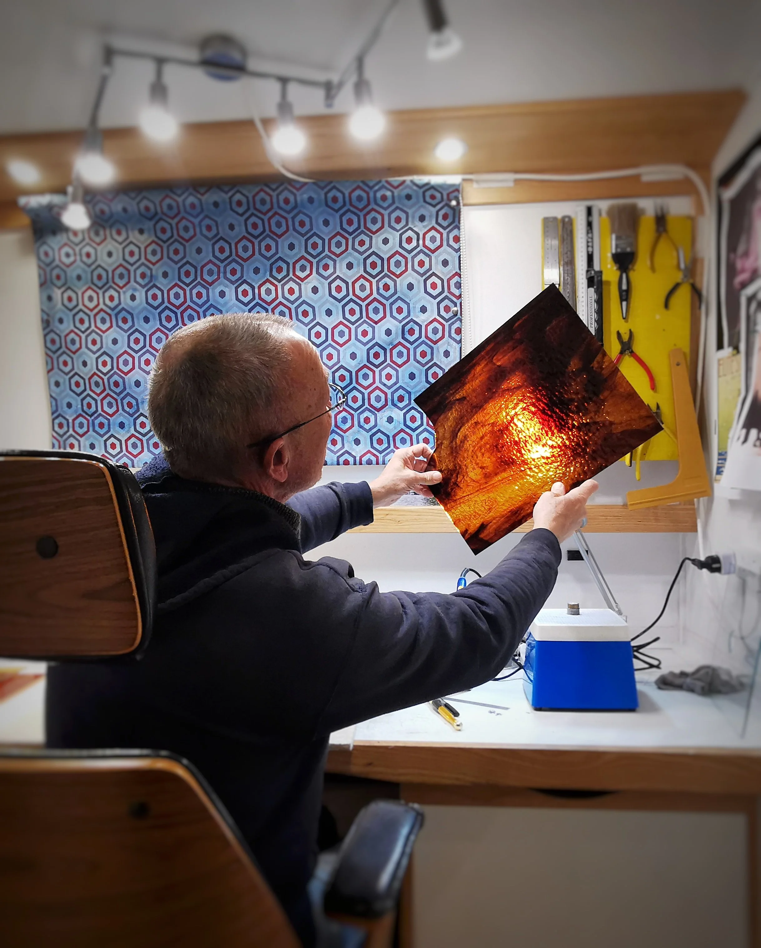 Person examining stained glass sheet at a workshop desk with tools and lighting overhead.
