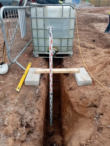 A construction site with a large water tank, a vertical measuring rod in the ground, and a narrow trench, steel barriers on the side, and tools nearby.