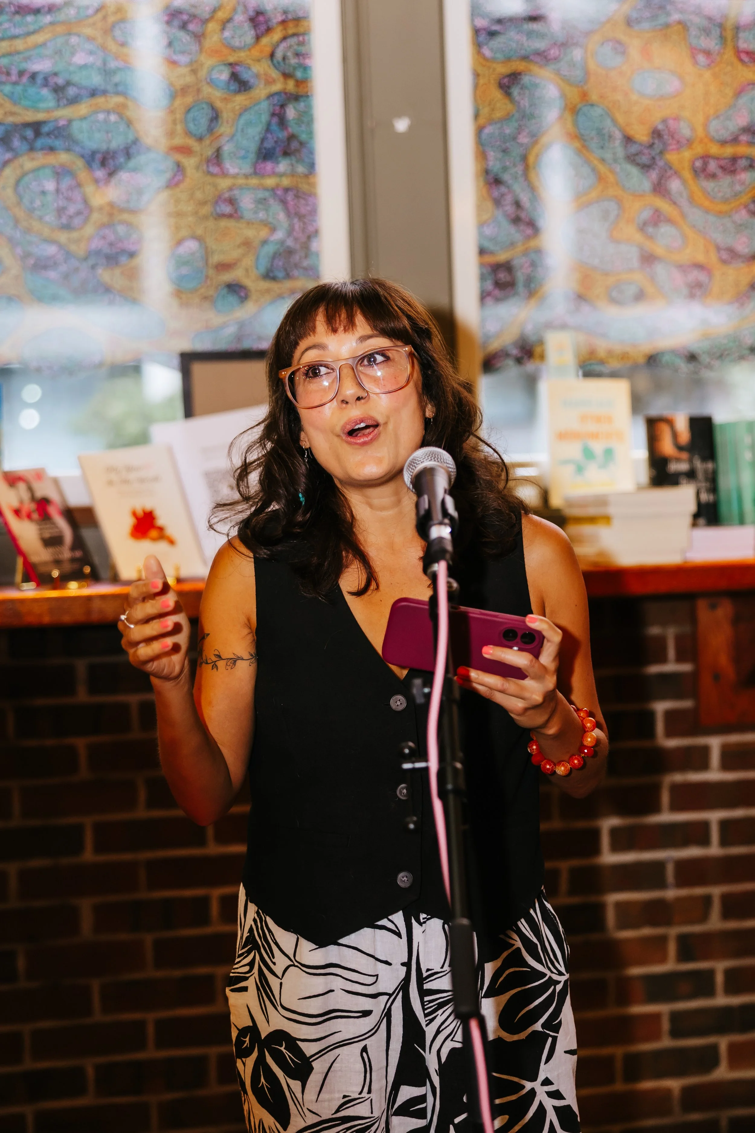 Woman with glasses speaking into a microphone, holding a tablet, standing in front of books and colorful abstract artwork.