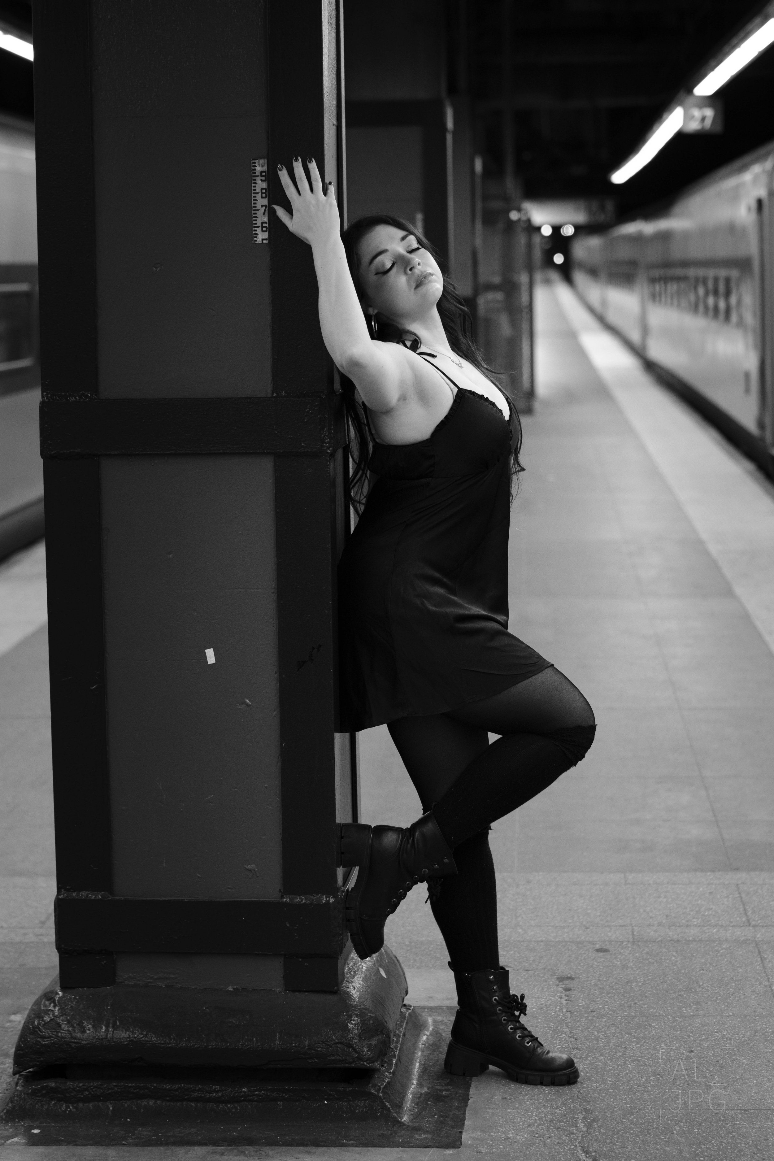 A woman in a black dress, tights, and boots standing against a platform in a subway station, with her eyes closed and arms raised, touching a support column.