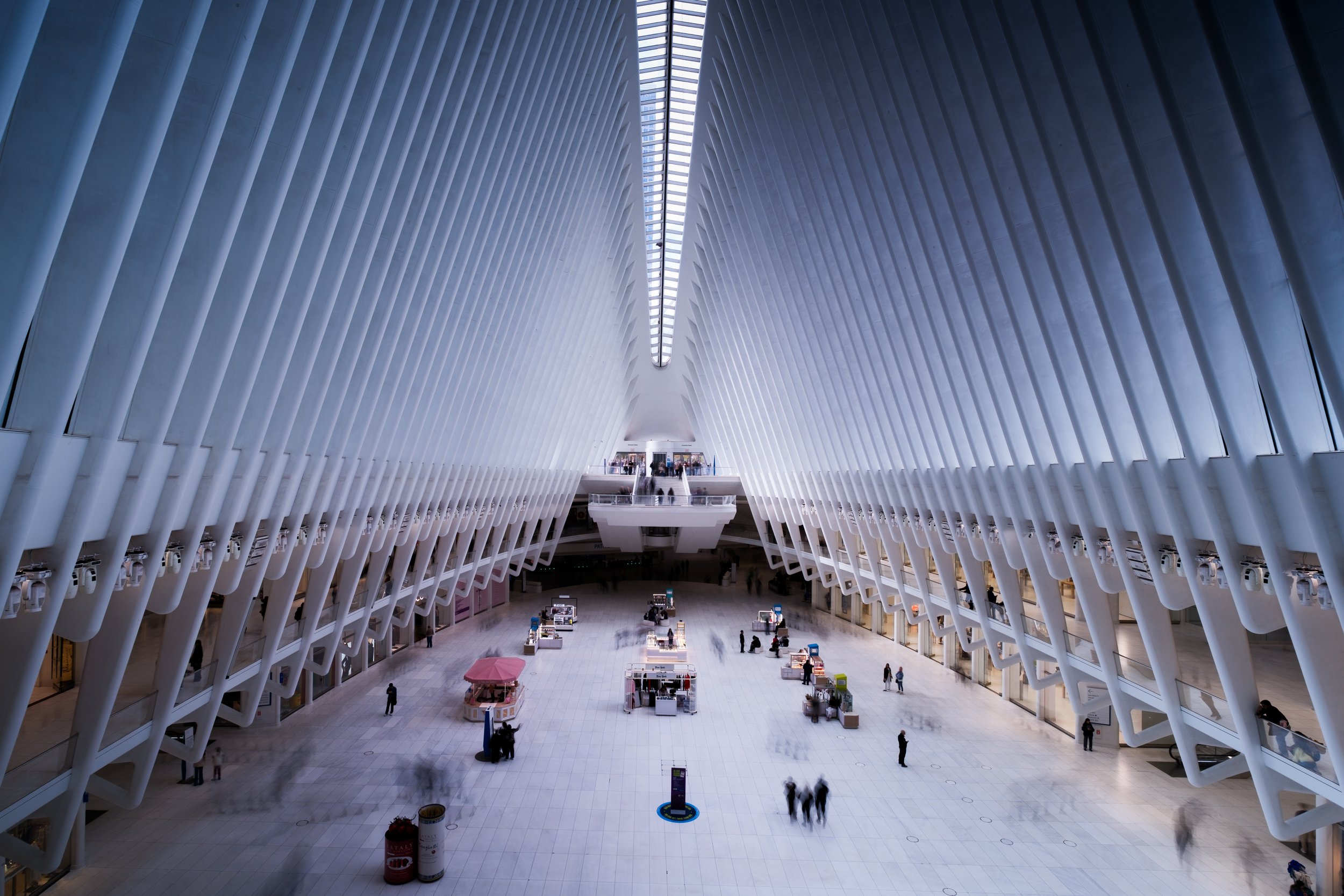 Interior of a modern, spacious airport terminal with high white architectural ceiling, scattered people, and small kiosk stalls on the floor.