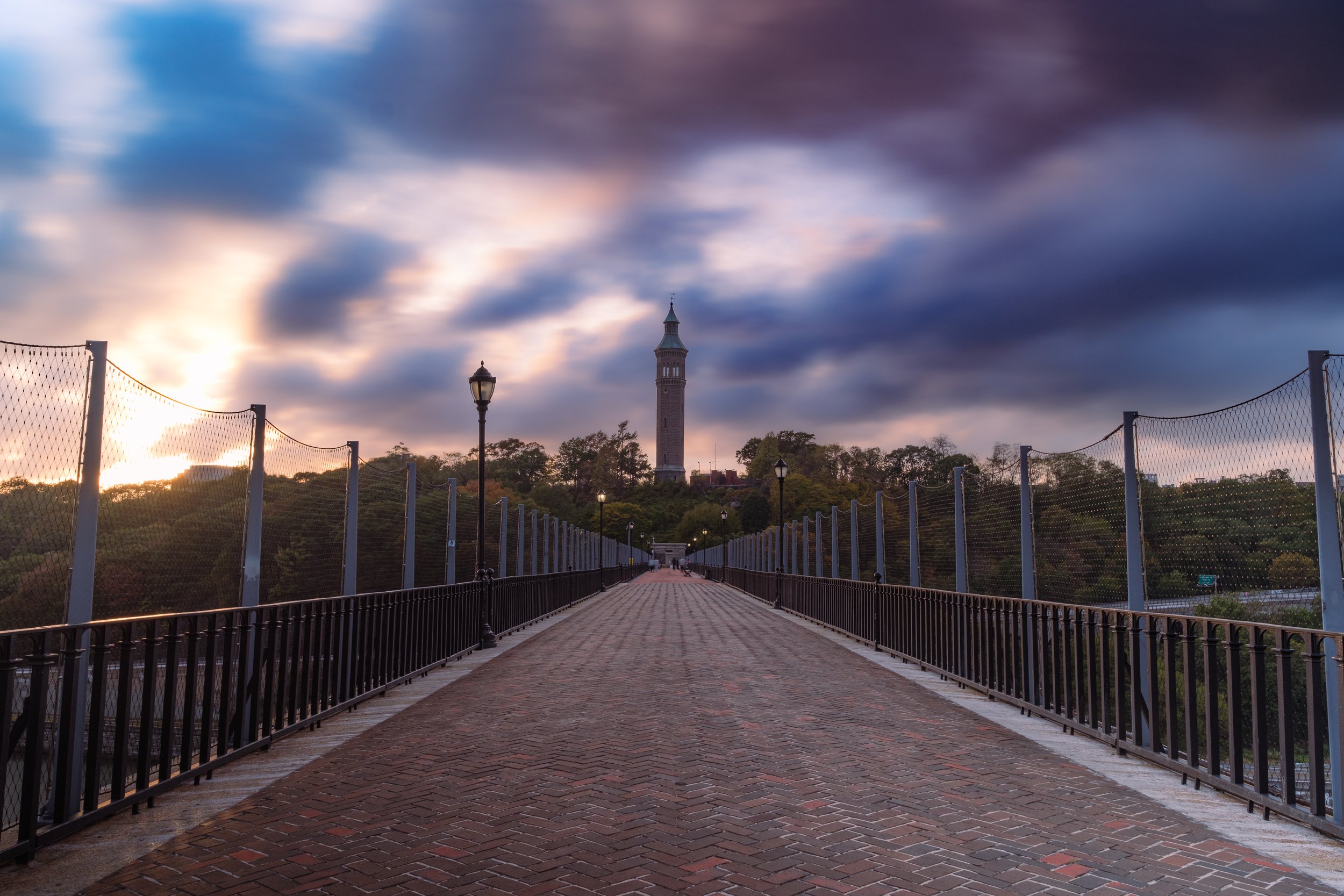 A brick pathway leading to a tall tower on a hill, with cloudy sky and sunset in the background.