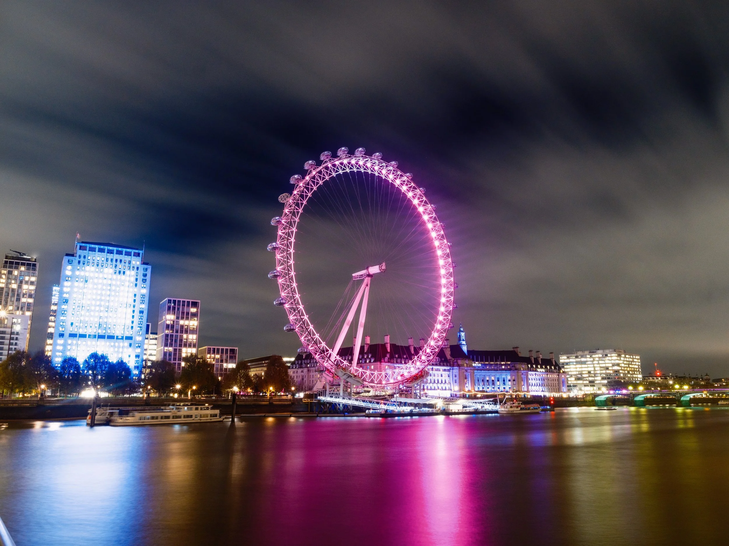 Nighttime view of the London Eye Ferris wheel illuminated in pink, reflecting on the River Thames, with city buildings lit up in the background.