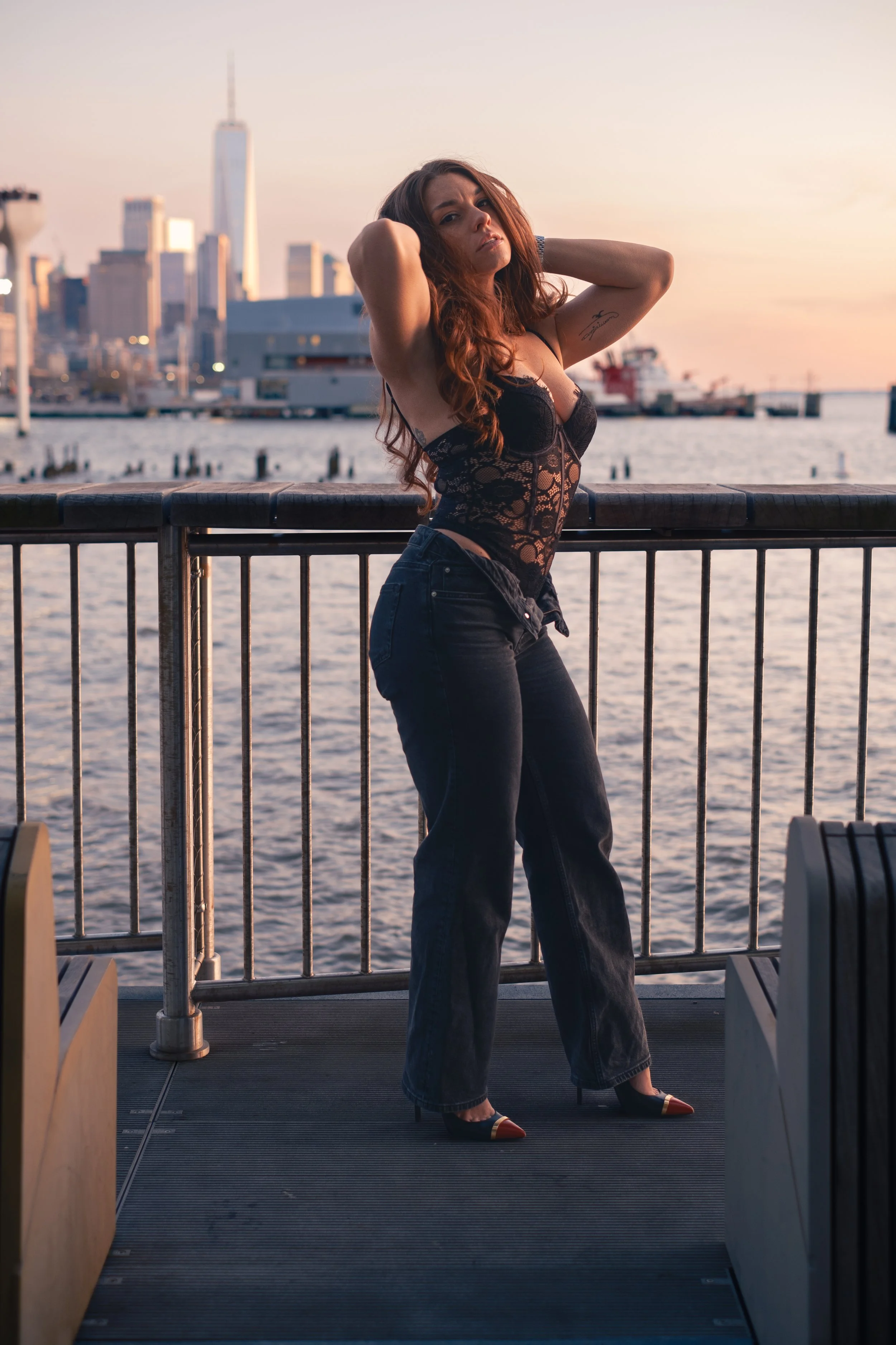 A woman with red hair poses on a waterfront at sunset, with a city skyline in the background, wearing a black lace top and black jeans.