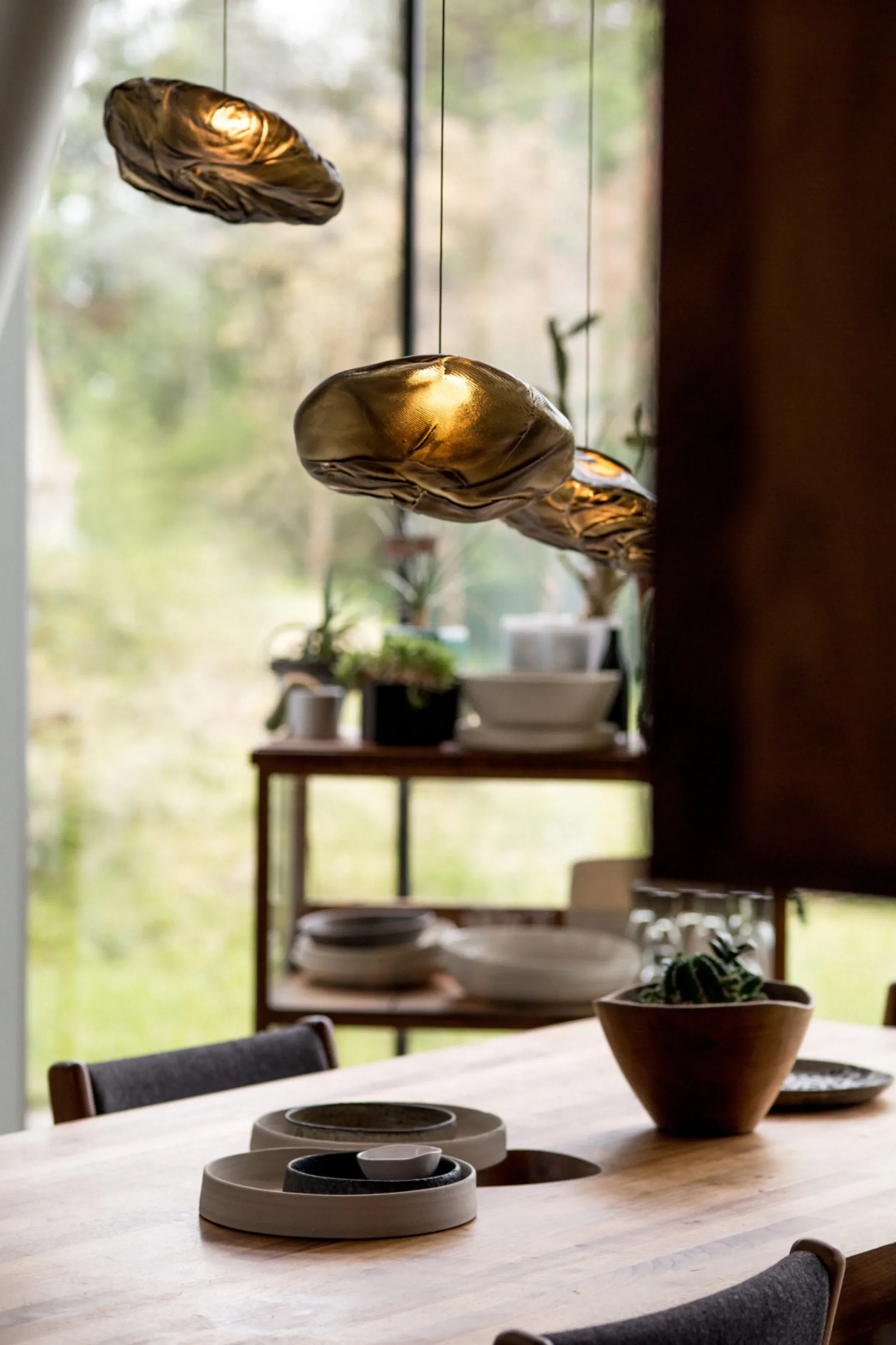 Decorative hanging metallic balloons over a wooden dining table, with a background of a window and shelves holding plates and potted plants.