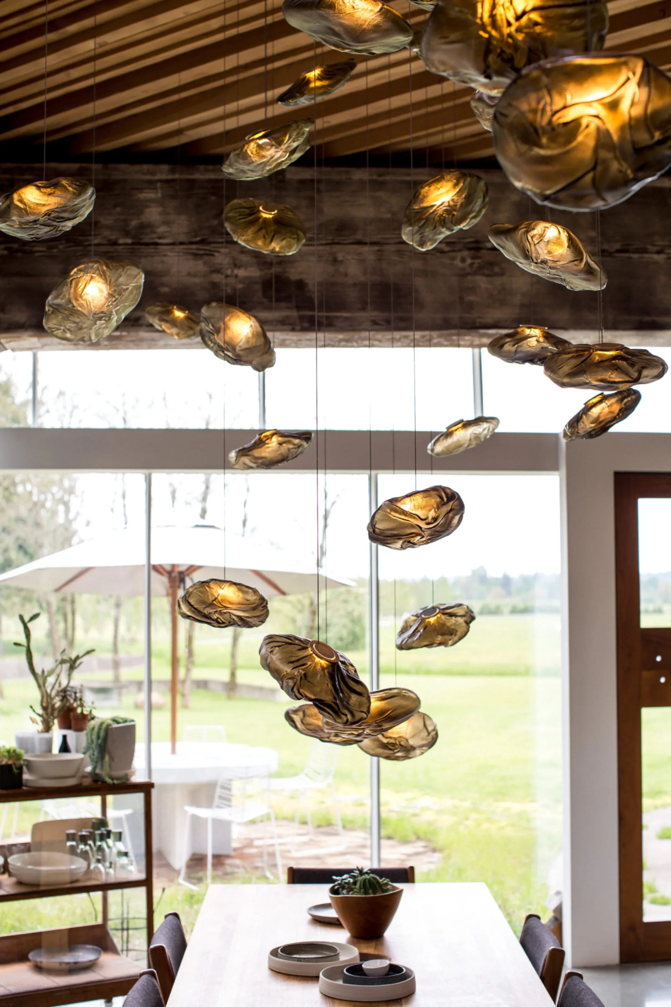 Modern hanging light fixtures made of crumpled metal shades hanging above a dining table in a bright room with large windows and a view of an outdoor patio with a white umbrella and outdoor furniture.