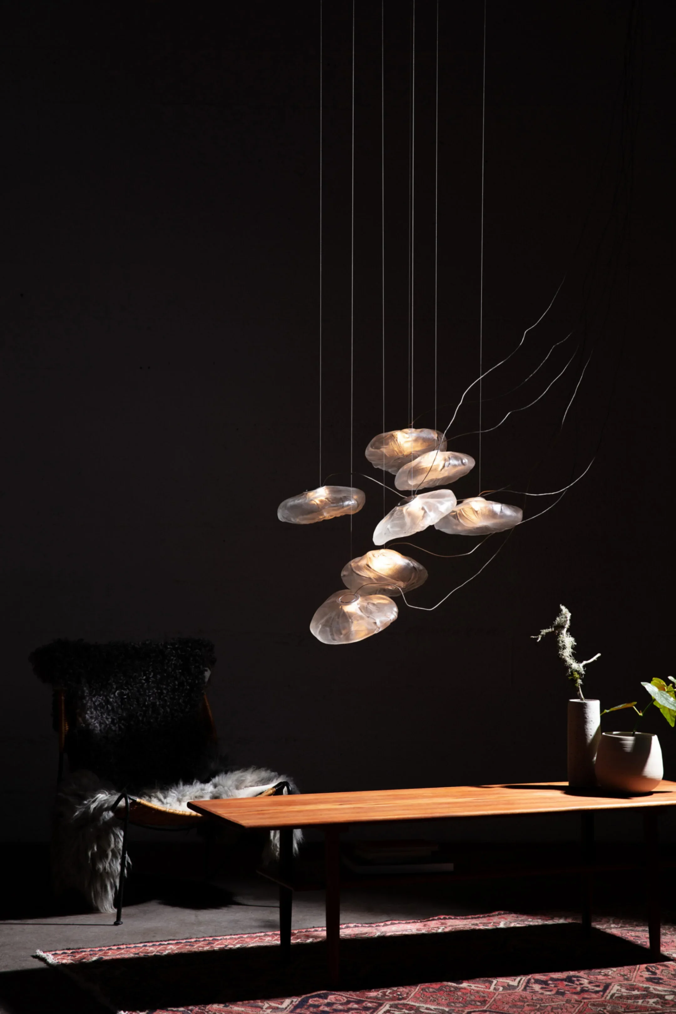 Modern wooden coffee table with two vases and a small plant, black chair with sheepskin throw, and hanging wall lamp with multiple glass shades, in a dimly lit room.