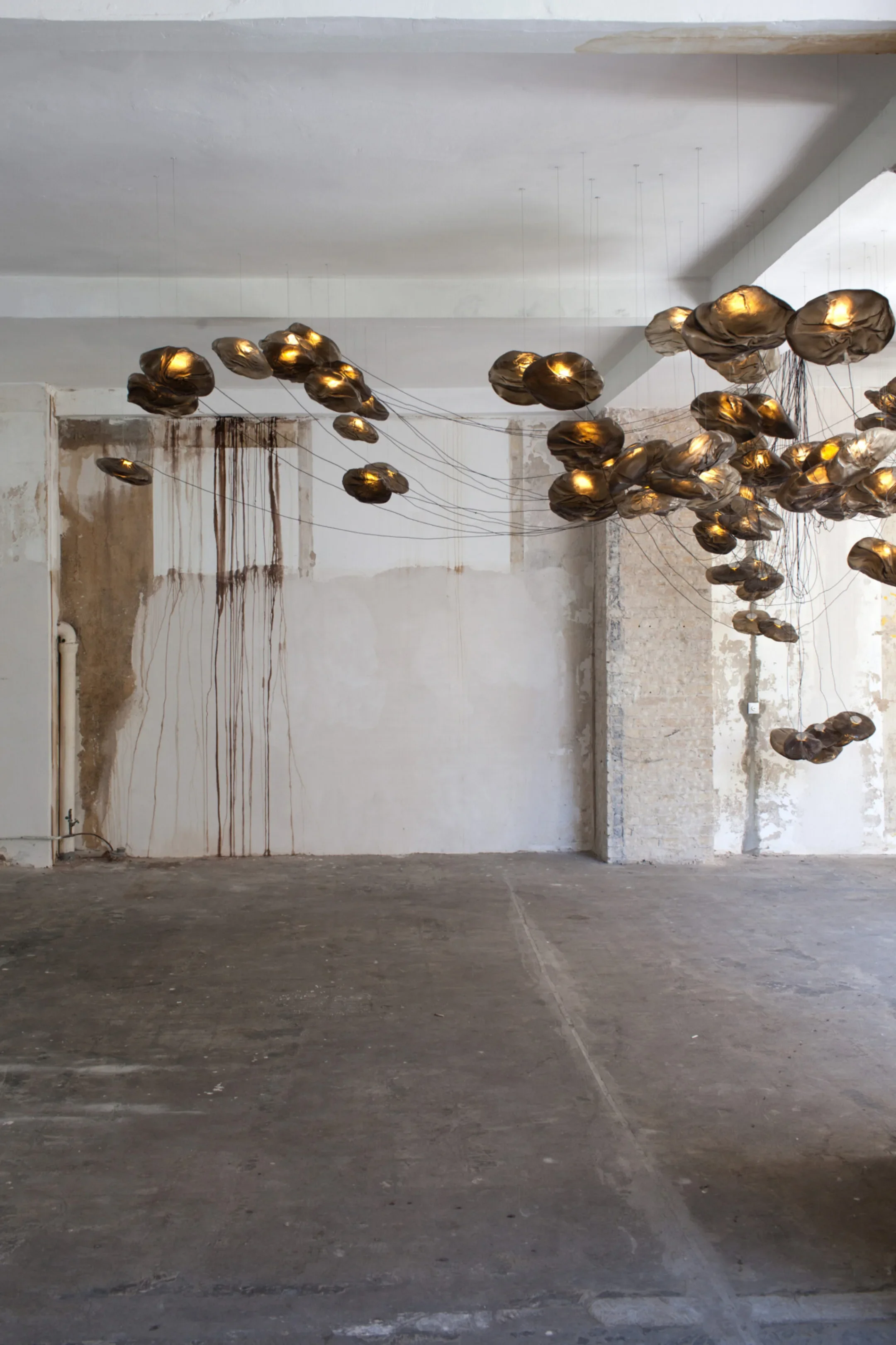 Art installation with clustered metallic balloons hanging from thin wires in an unfinished room with exposed concrete walls and floor.