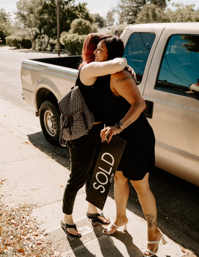 Two women hugging on a sidewalk, one holding a  sold sign, near a pickup truck.