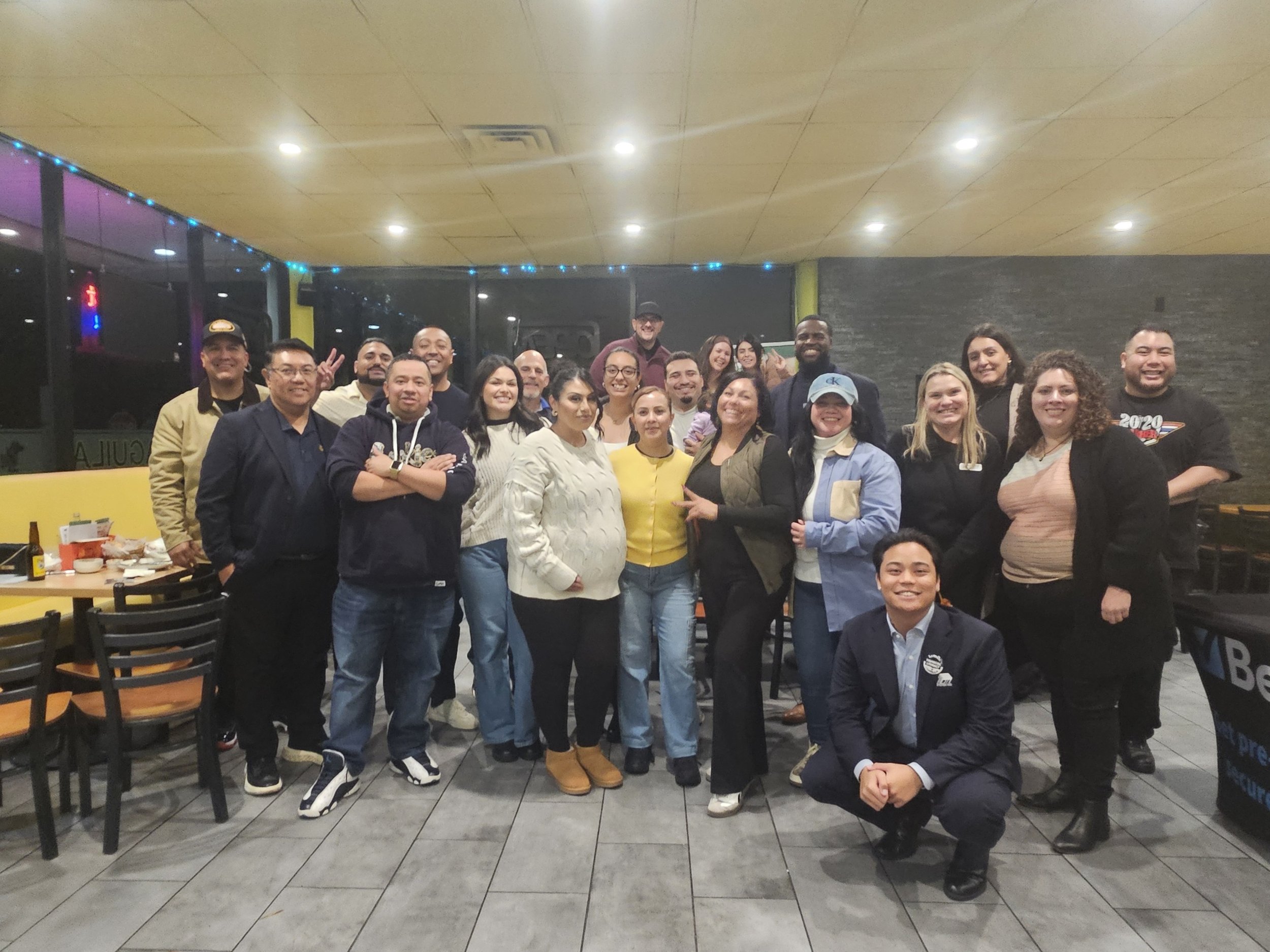 Group of smiling people gathered inside a restaurant, some standing and one squatting in front, celebrating or attending an event.