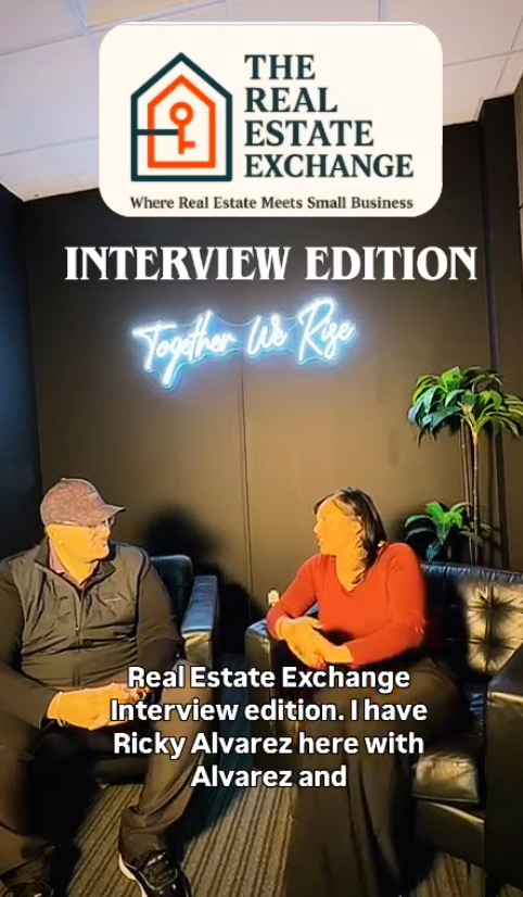 Two people seated on black leather chairs in a lounge area, engaged in conversation. The man wears a gray hat and dark clothing, and the woman wears a red top. A neon sign behind them reads "Together We Rise," and there is a large potted plant on the