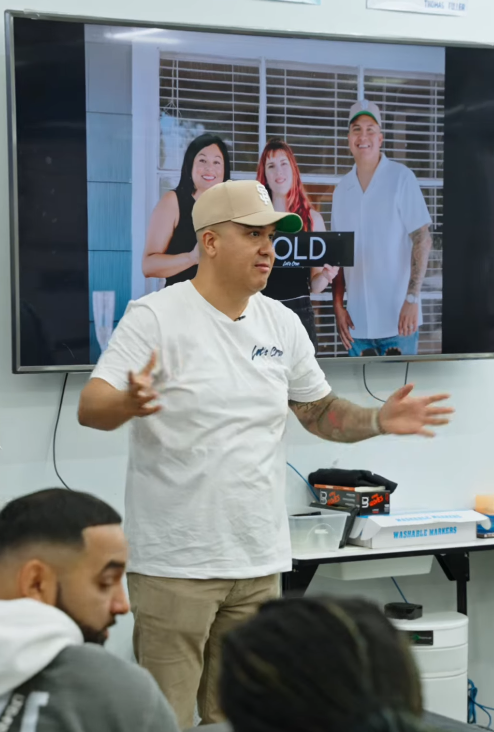 A man wearing a beige cap and white t-shirt speaking in front of a group, with a large screen behind him showing a photo of three people smiling, one woman with black hair, a woman with red hair, and a man in a white shirt.