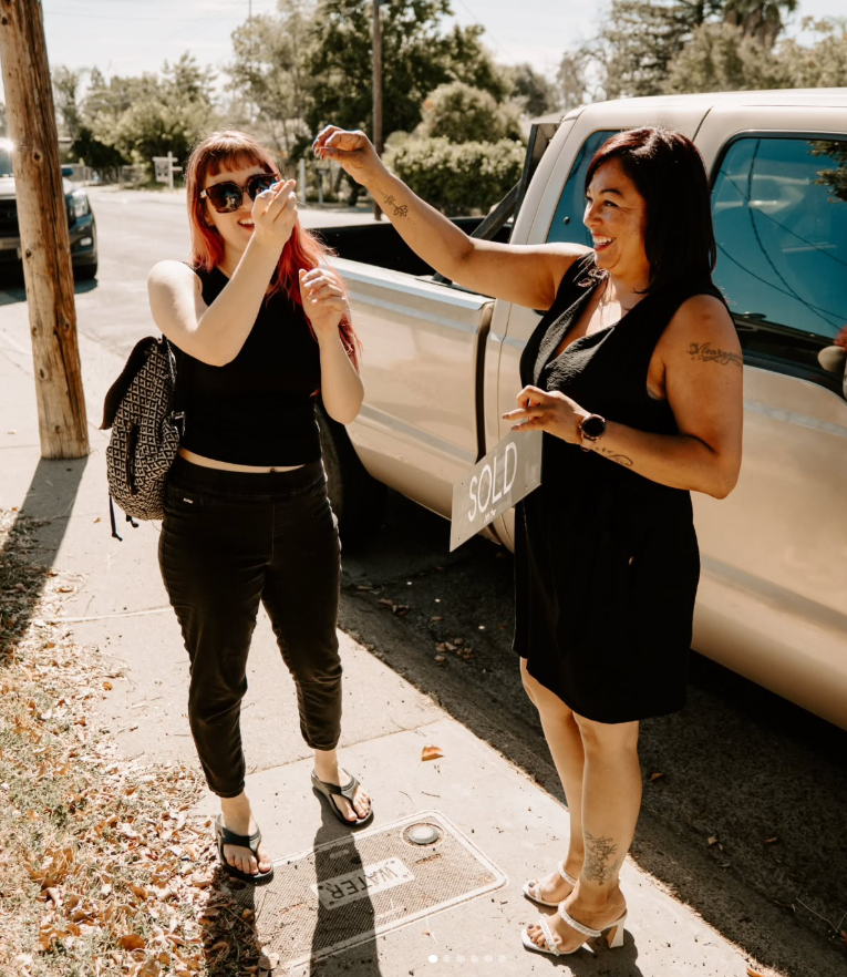 Two women standing on a sidewalk next to a pickup truck, one holding a sold sign, smiling and celebrating a car purchase.