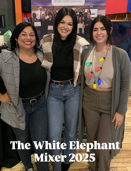 Three women smiling and posing together at the White Elephant Mixer 2025 event, with a group photo and event sign in the background.