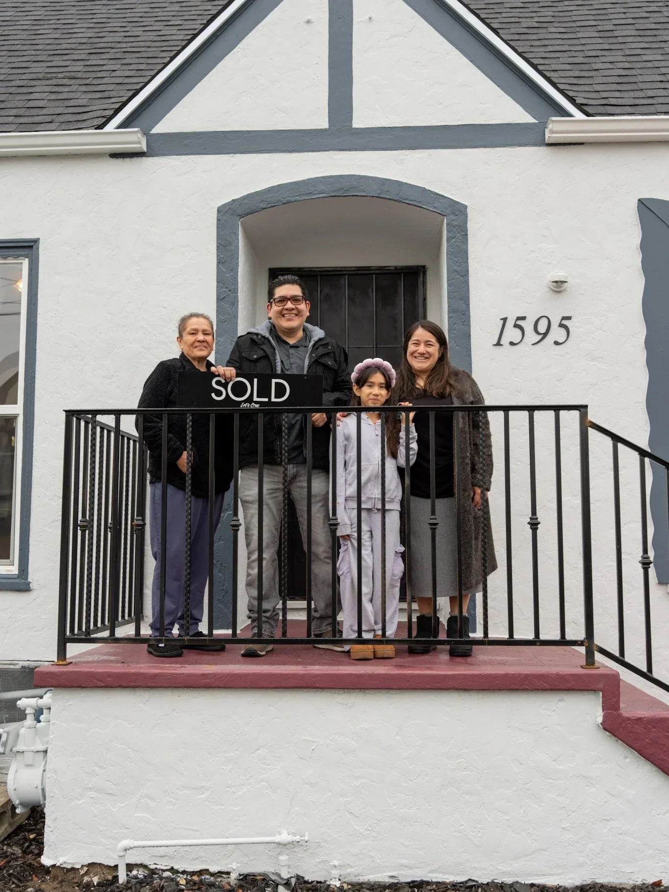 A family of four standing on the front porch of a house that is marked as sold, with a sign that says 'SOLD' in front of the door, all smiling. The house is white with dark trim and the number 1595 displays beside the door.