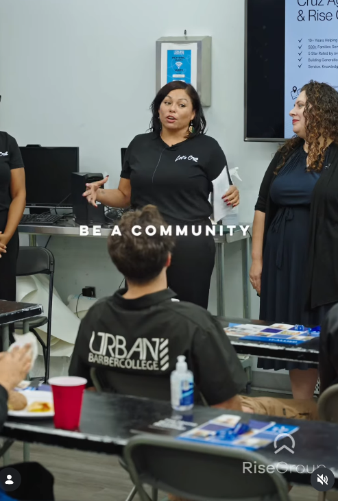 Two women speaking to a group in a classroom or meeting room setting, with one woman holding papers. Several people are seated at tables with brochures, one person wears a 'Urban Barber College' shirt, and there is a large screen or monitor on the wa